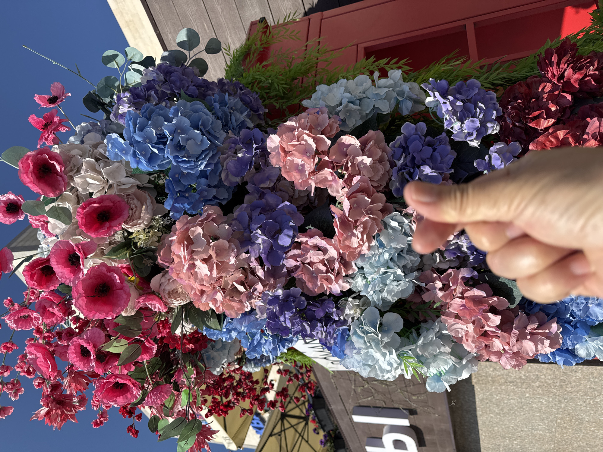 A close-up, low-angle shot captures an abundant floral arrangement in Kokshetau, Kazakhstan, against a clear blue sky. The arrangement features a variety of artificial flowers, predominantly hydrangeas and poppies, in vibrant shades of pink, red, blue, purple, and light blue. The flowers are densely packed, forming a large, colorful column. In the foreground, a human hand is performing the "finger heart" gesture, with the thumb and index finger touching to form a heart shape. Behind the floral arrangement, elements of an urban environment are visible, including a building facade with brown wooden paneling and a sign with white lettering that appears to read "Ы" followed by other obscured characters. A red structure, possibly a door or partition, is also visible on the right side of the image. The scene suggests an outdoor setting, possibly a decorative display outside a shop or restaurant.