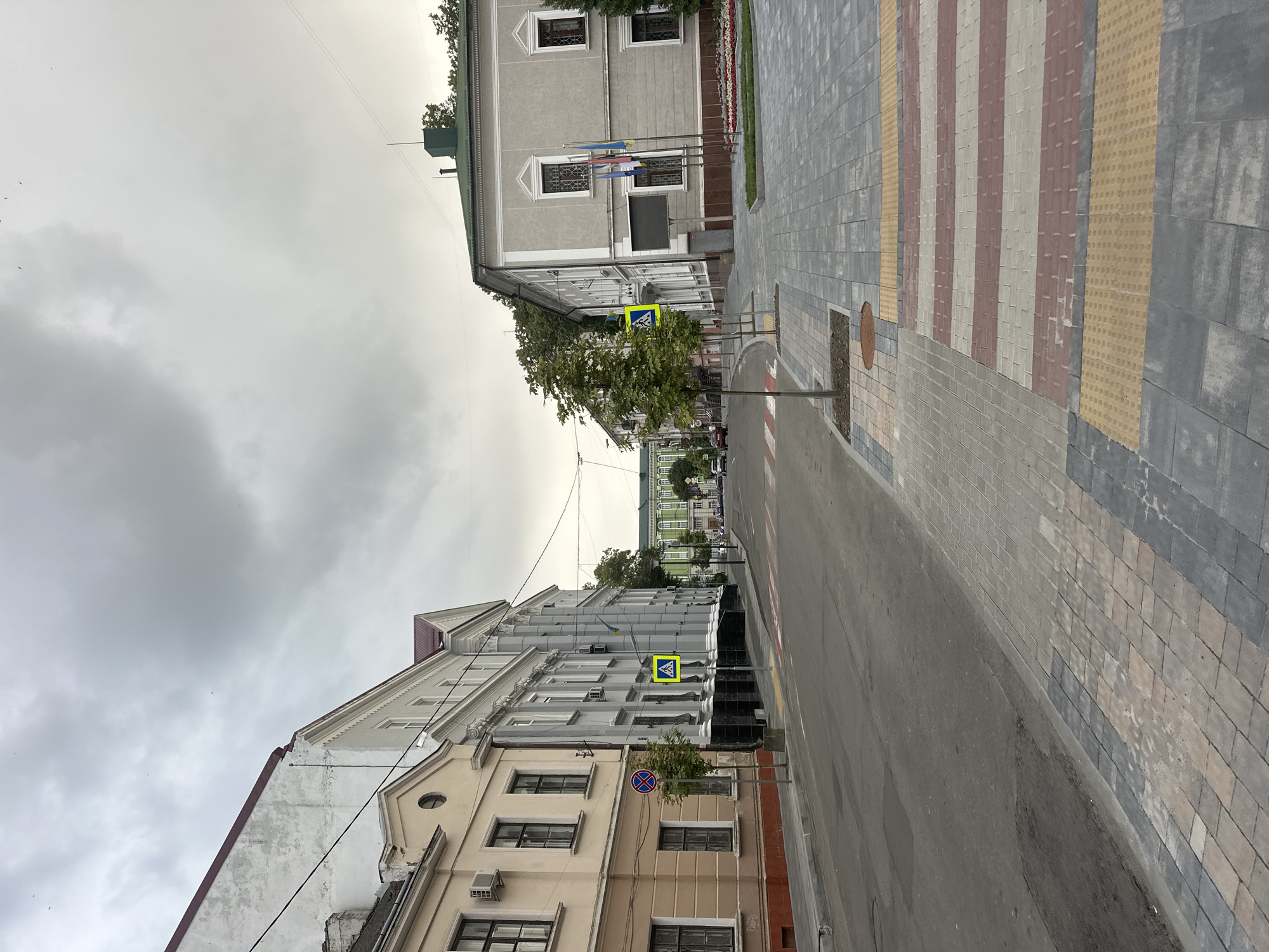 The image shows a street scene in Ternopil, Ukraine, likely in the city center given the architecture and paving. On the left, a light beige building with dark windows and a dark red roof is visible, followed by a larger grey building with multiple windows and classical architectural elements. On the right, a light grey building with a green roof and several windows stands next to a modern paved sidewalk. The sidewalk features a pedestrian crossing with red and white striped paving, bordered by tactile paving. Trees line the sidewalk. The street is mostly empty, with a few cars visible in the distance. The sky is overcast and grey, suggesting a cloudy day. The scene appears to be a quiet urban environment, possibly in the morning or late afternoon due to the soft lighting.