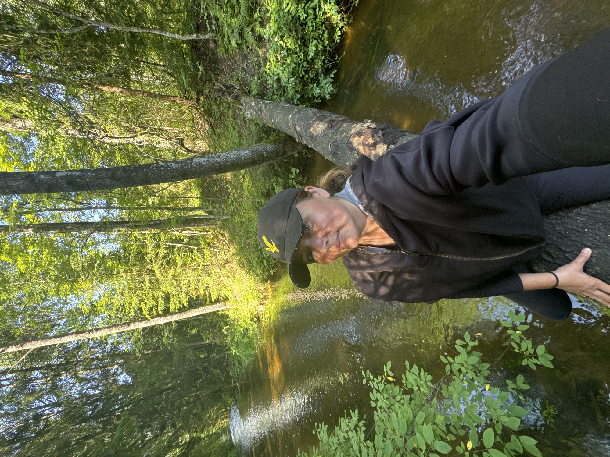 A woman is captured in a selfie on a fallen log bridging a small river in a lush, sun-dappled forest. She is wearing a black baseball cap with a yellow emblem and a black zip-up hoodie over a grey layer. Her facial expression is neutral as she looks towards the camera. The forest canopy is dense with green foliage, and sunlight filters through the leaves, illuminating the water and the forest floor. The river is shallow and clear, with a gentle current. The overall setting suggests a serene, natural environment within the city of Zelonaya Dolina, Russia.
