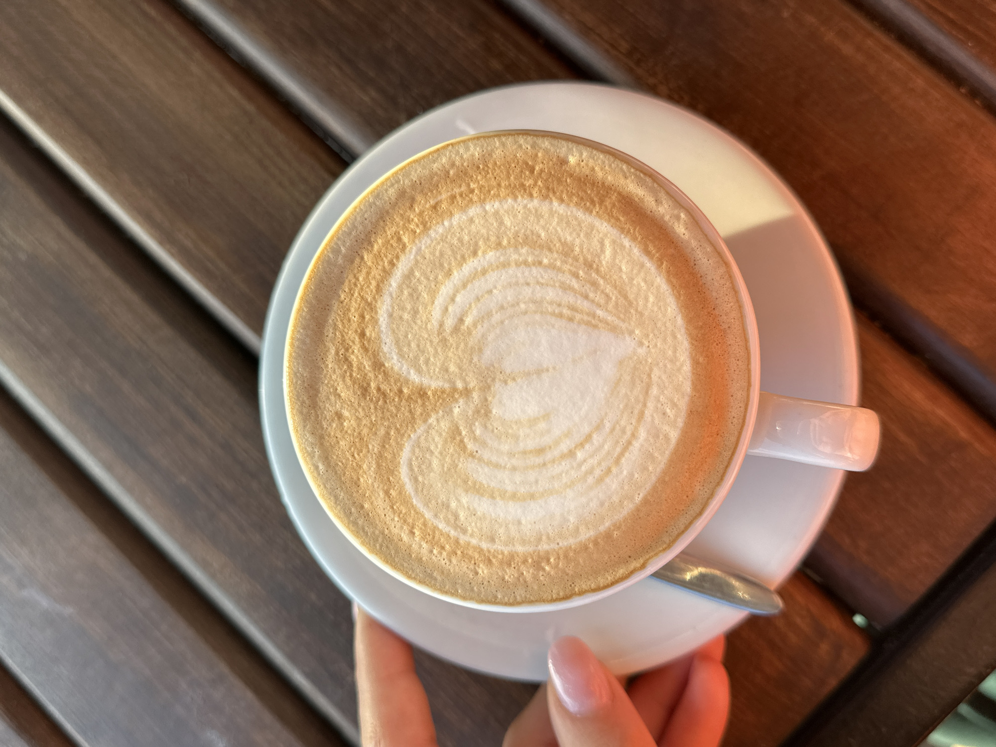 A top-down shot shows a white coffee cup, filled with a latte featuring a heart-shaped latte art design, resting on a white saucer. A spoon rests near the saucer's edge. A person's hand with a pink manicure is visible holding the saucer. The cup and saucer sit on a wooden surface consisting of parallel planks. The image was likely taken outdoors in Šušanj, Montenegro, based on the available context.