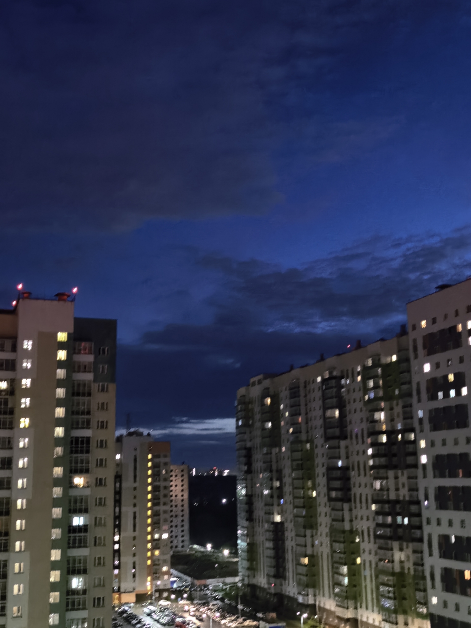 The image is a nighttime exterior view of a residential area. In the foreground, two tall apartment buildings are prominently featured. The building on the left is lighter in color with visible windows, some of which are illuminated. It has red lights on its rooftop. The building on the right is also a multi-story apartment building with numerous illuminated windows, displaying a mix of white and green on its facade. In the background, a third, slightly shorter apartment building can be seen, with its windows also lit. Beyond the buildings, a city skyline with distant lights is visible against a dark, cloudy sky. The lower portion of the image shows a parking lot filled with cars. The scene depicts a typical urban environment at dusk or night.