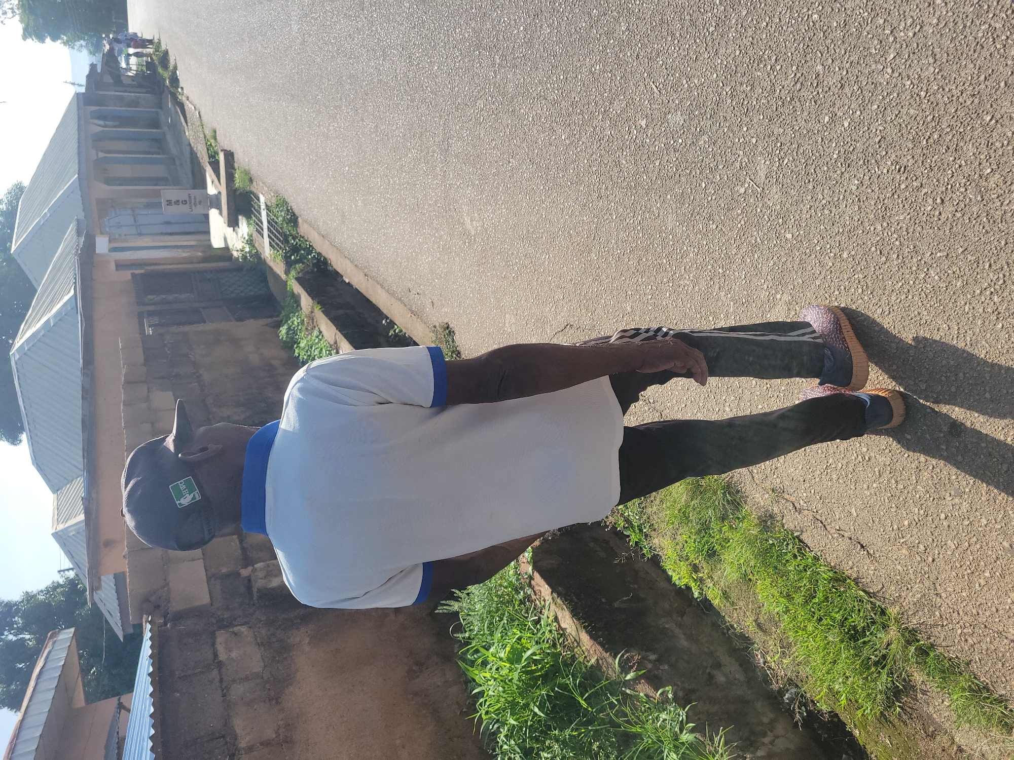 In Jos, Nigeria, a person is captured from behind, walking on a paved road. The individual is wearing a white polo shirt with blue trim, dark pants with white stripes, and colorful sneakers. A dark baseball cap with a green and white emblem, possibly the NBA logo, is on their head. The person is walking away from the camera, towards buildings lining the street. These structures have light-colored walls and corrugated metal roofs, with some featuring open-air covered walkways. Green vegetation grows along the side of the road and in front of the buildings. A drainage ditch runs along the side of the road, adjacent to the vegetation. The road is asphalt and appears well-maintained, with clear sunlight casting shadows. The overall environment suggests a residential or commercial street in a built-up area of the city.
