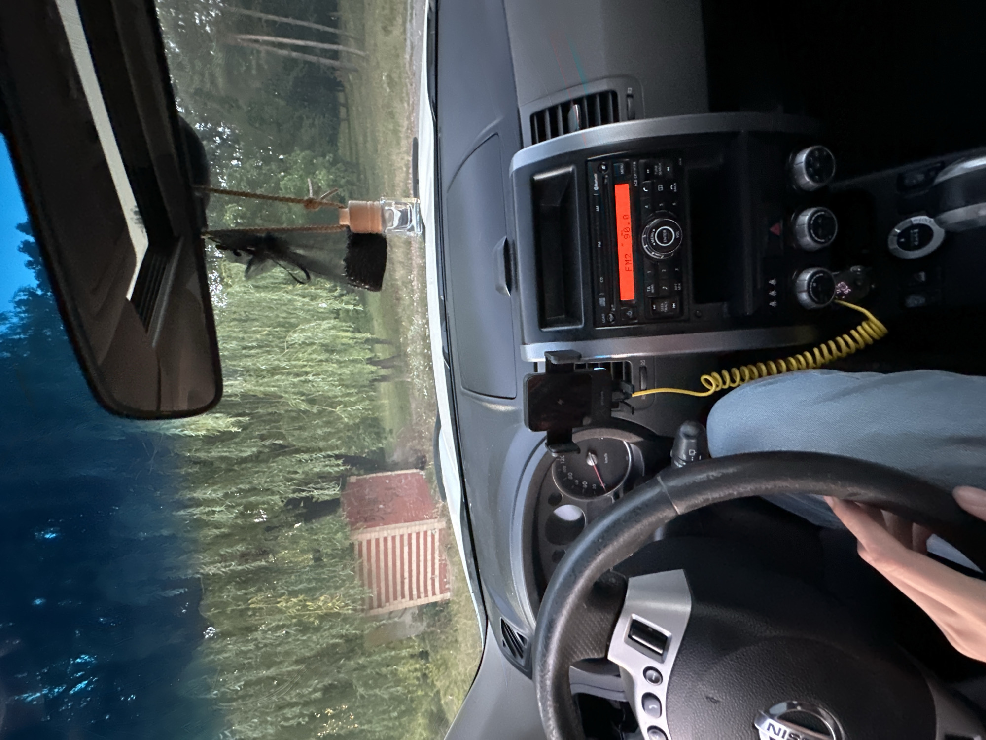 The image shows the interior of a car, likely a Nissan, with a driver's hand resting on the steering wheel. The dashboard features a car radio displaying "FM2 90.0", a phone mounted on a car holder, and a yellow coiled cable. Outside the car, through the windshield, a backdrop of trees and a red and white striped structure is visible. This scene is situated in Makiv, Ukraine, as indicated by the metadata.
