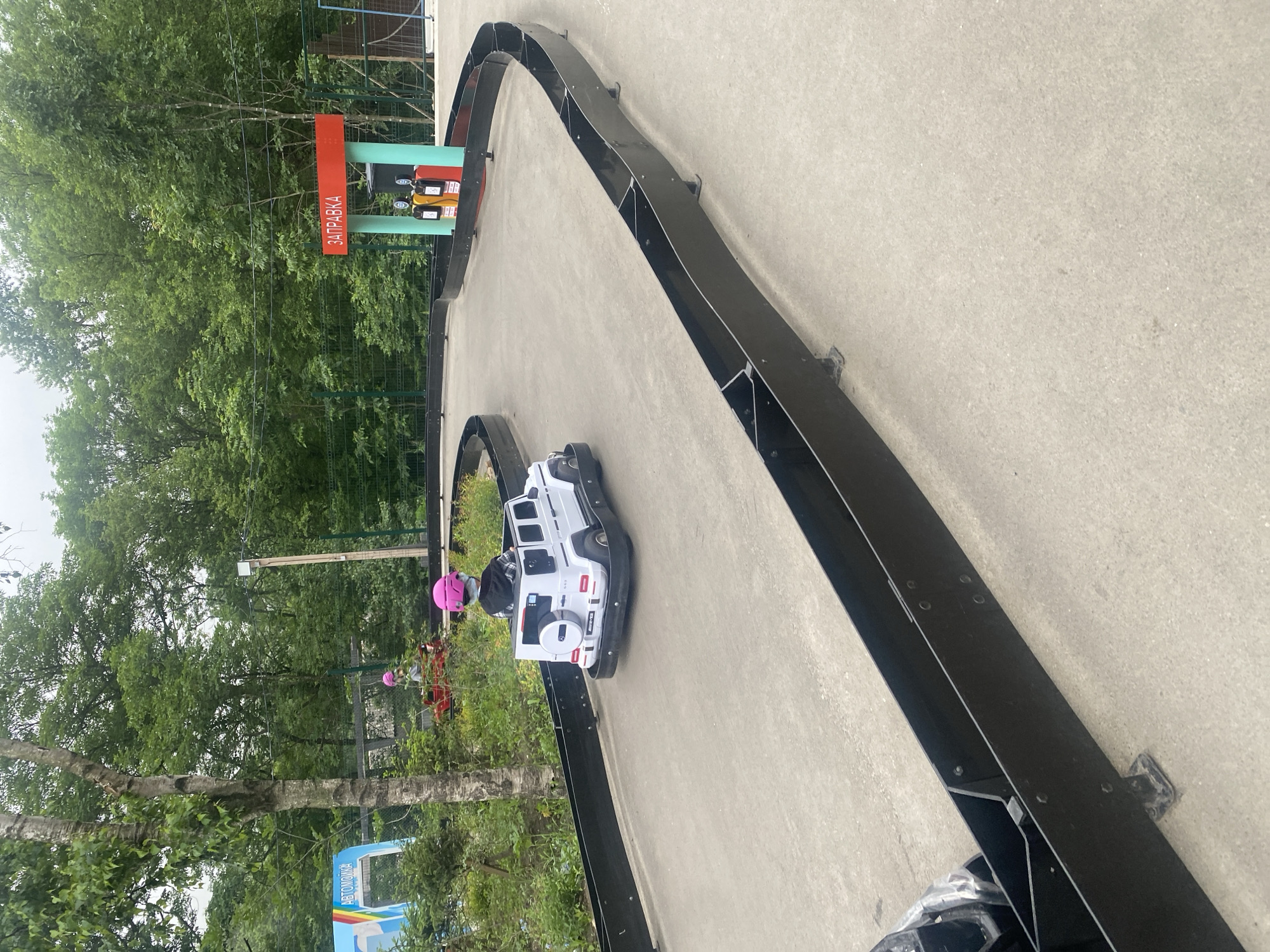 In an outdoor amusement park, a young child wearing a pink helmet navigates a white toy car along a go-kart track. The track, bordered by black plastic barriers, curves around a paved area. In the background, a miniature gas station, labeled "ЗАПРАВКА" (which translates to "GAS STATION"), stands with two visible gas pumps. Lush green trees form a dense backdrop, suggesting a location surrounded by nature. The sky is overcast, indicating a cloudy day.