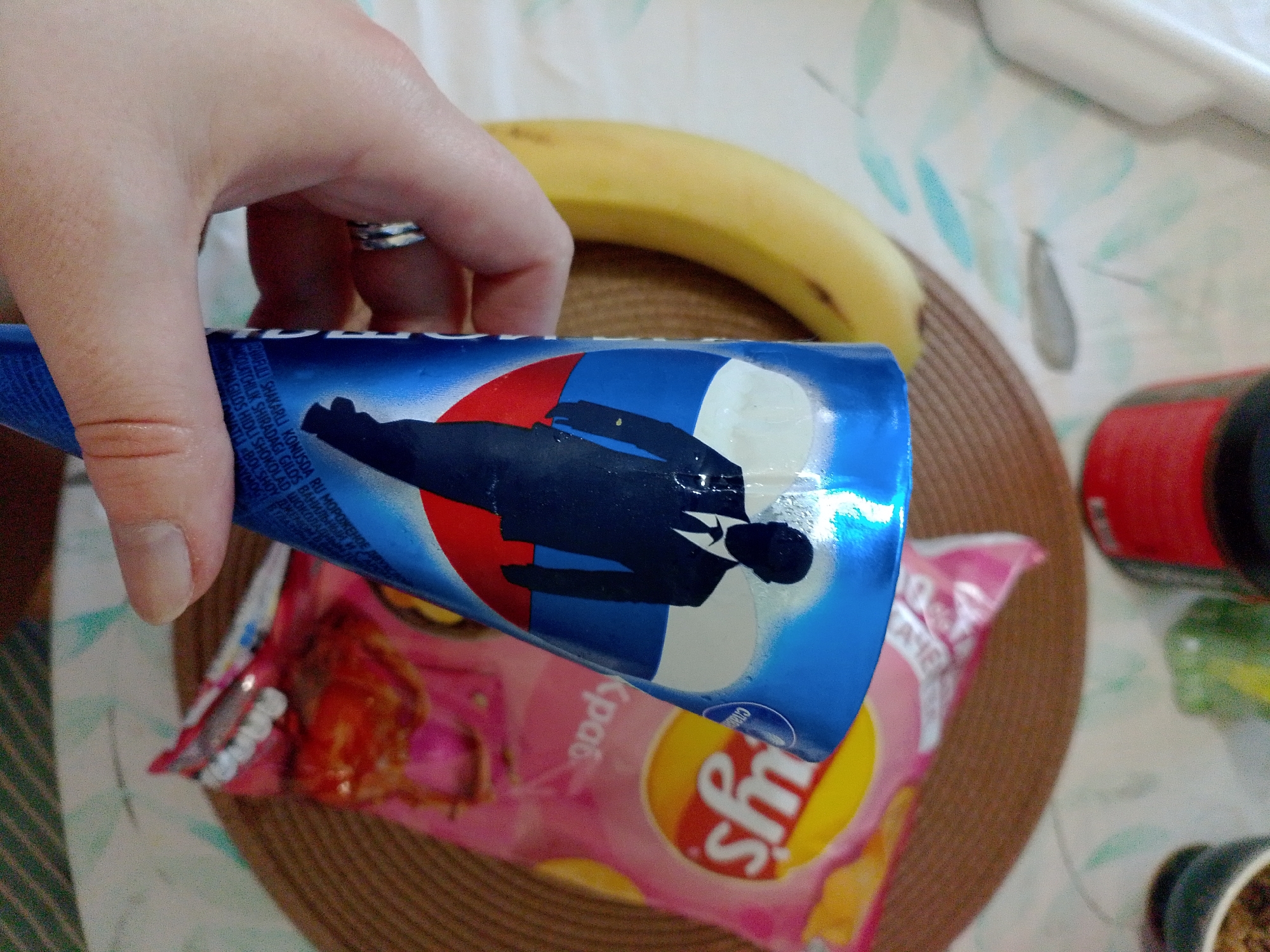 A hand is holding a blue Cornetto ice cream wrapper in front of a table. The wrapper features a stylized illustration of a man in a black suit against a red, white, and blue background. Underneath the wrapper is a bag of Lay's crab chips. Behind the wrapper, there is a banana and a brown placemat. Other items on the table include a red container and a small plant in a pot. The image does not provide specific information about the location, city, or country.