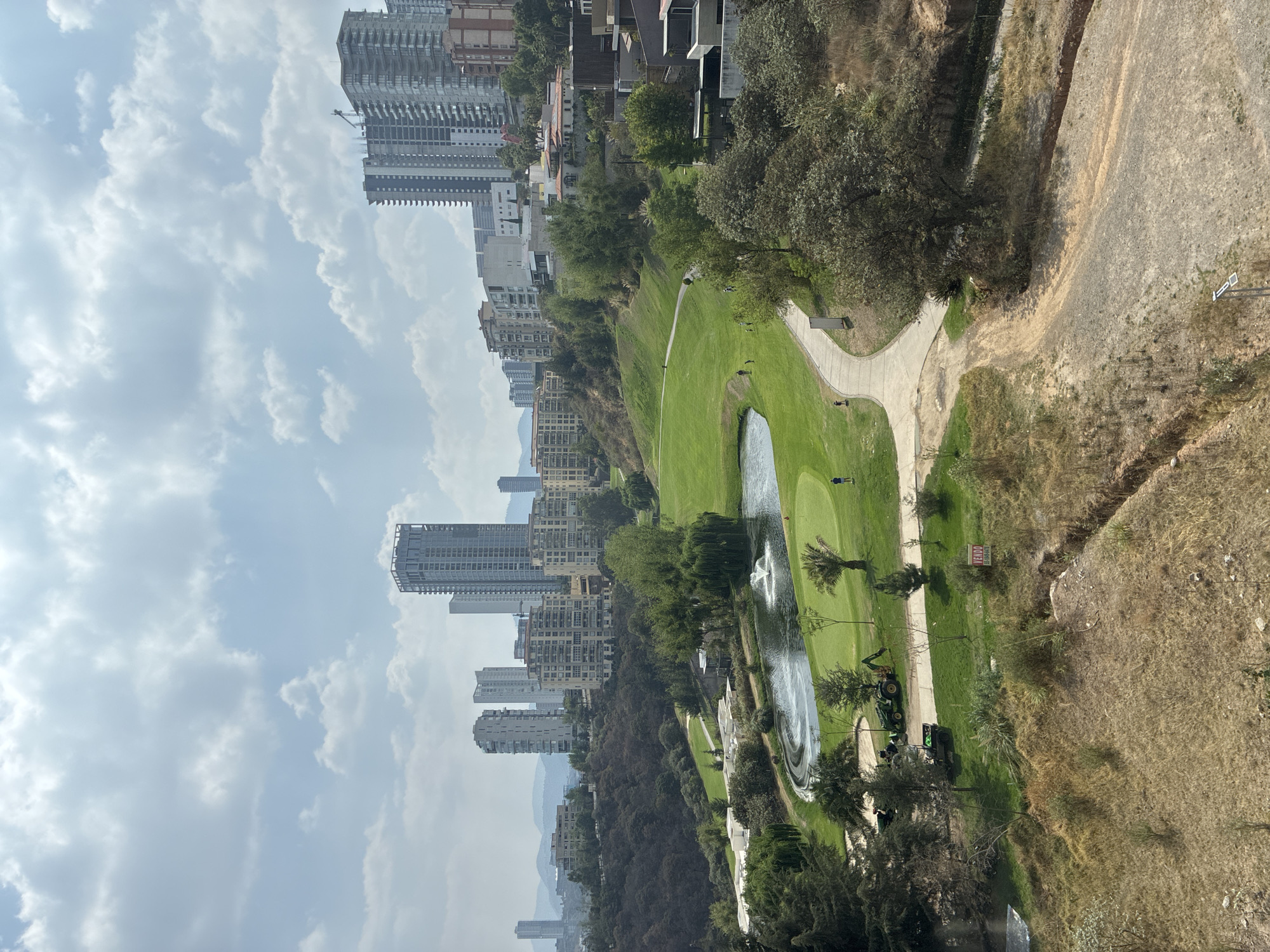 The image presents an elevated view of a green landscape, identified as a golf course, adjacent to dense urban development in Huixquilucan de Degollado, Mexico. The golf course features a water feature with a fountain. People are visible on the golf course. Golf maintenance vehicles are on the edge of the golf course. A beige hillside extends to the right of the golf course. High-rise buildings and residential structures dominate the skyline to the left of the course. The sky is cloudy.
