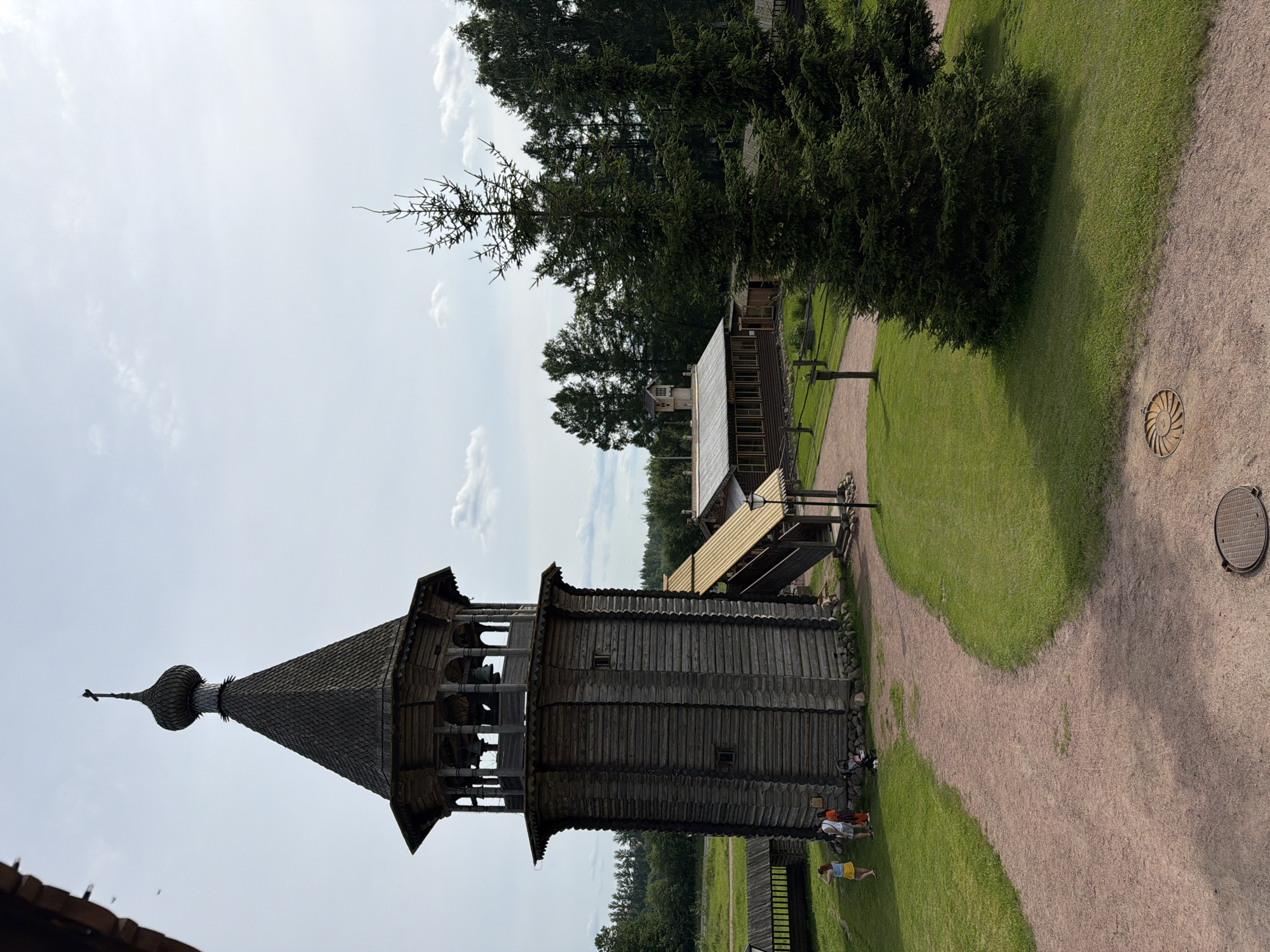 The image depicts an outdoor scene in the city of Imeni Sverdlova, Russia, featuring a traditional wooden bell tower with a cross on its roof. Adjacent to the bell tower is a long, low-rise wooden building with a corrugated roof, and further in the background, a dense line of evergreen trees forms the horizon. A winding dirt path cuts through a well-maintained grassy area, where a few individuals, including two women and a person with a stroller, are visible near the base of the bell tower. The sky is partly cloudy, indicating fair weather. In the foreground, there are two circular metal grates embedded in the ground.