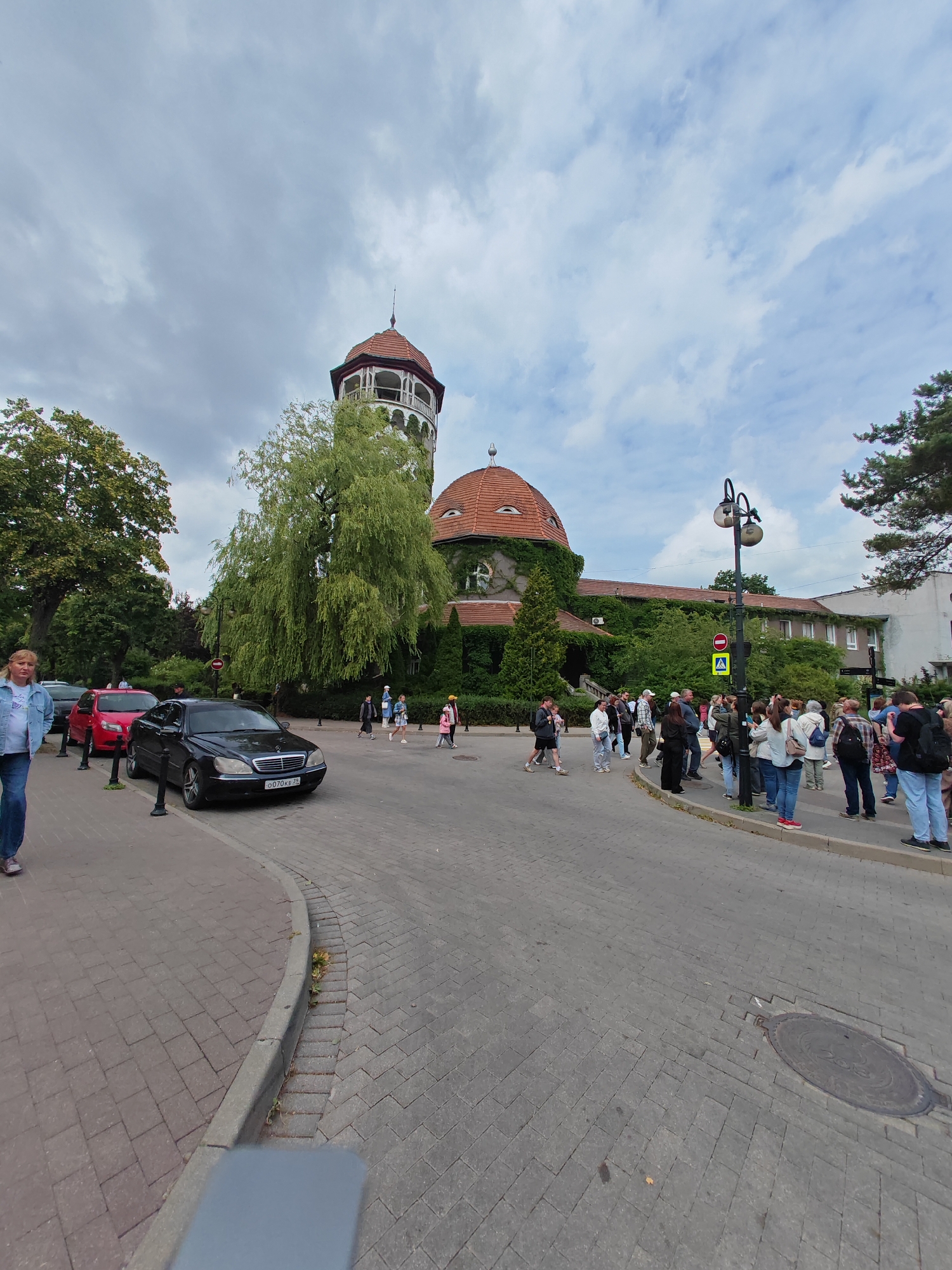 The image depicts a street scene in Svetlogorsk, Russia, on a cloudy day. A group of people are gathered, some walking and some standing, on a paved walkway. In the background, a distinctive building with a red tiled roof and a tower with white decorative railings is visible, partially covered by lush green ivy. A large, weeping willow tree stands prominently in front of the building. Several cars are parked along the side of the street, including a black Mercedes-Benz with a license plate that reads "O070KE 77". A woman in a denim jacket and jeans is walking on the left side of the foreground. A street lamp with two lights stands near the center of the image, accompanied by traffic signs indicating caution and a prohibition. The overall atmosphere appears to be that of a public gathering or a tourist area.
