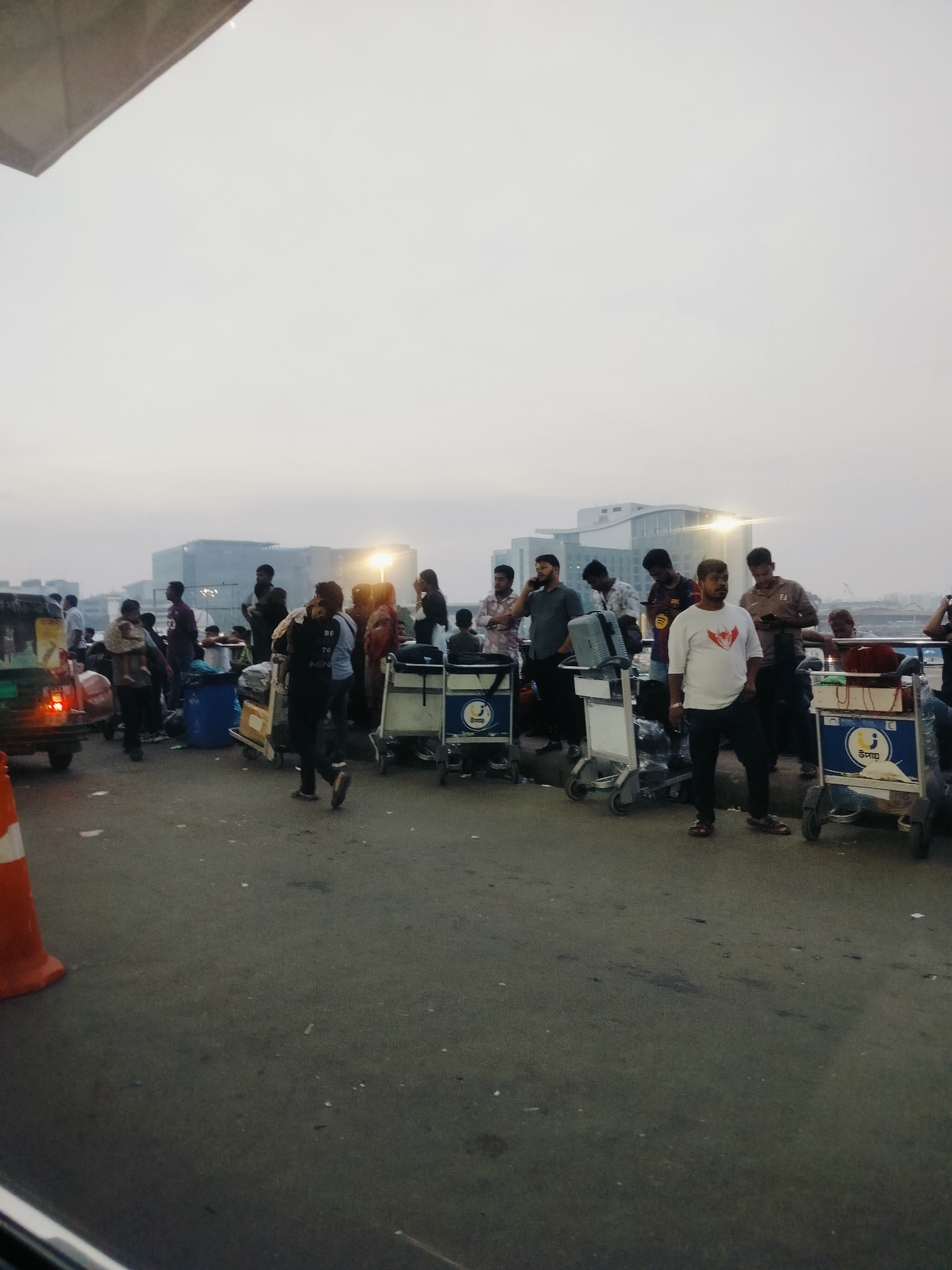 The image depicts a scene at Dhaka airport, Bangladesh, likely in the early morning or late evening due to the soft lighting. A group of people are gathered with luggage carts, suggesting they are either arriving or departing. A rickshaw is visible on the left, indicating local transportation. In the background, modern airport buildings with illuminated windows are present. The overall atmosphere is one of transit and movement.