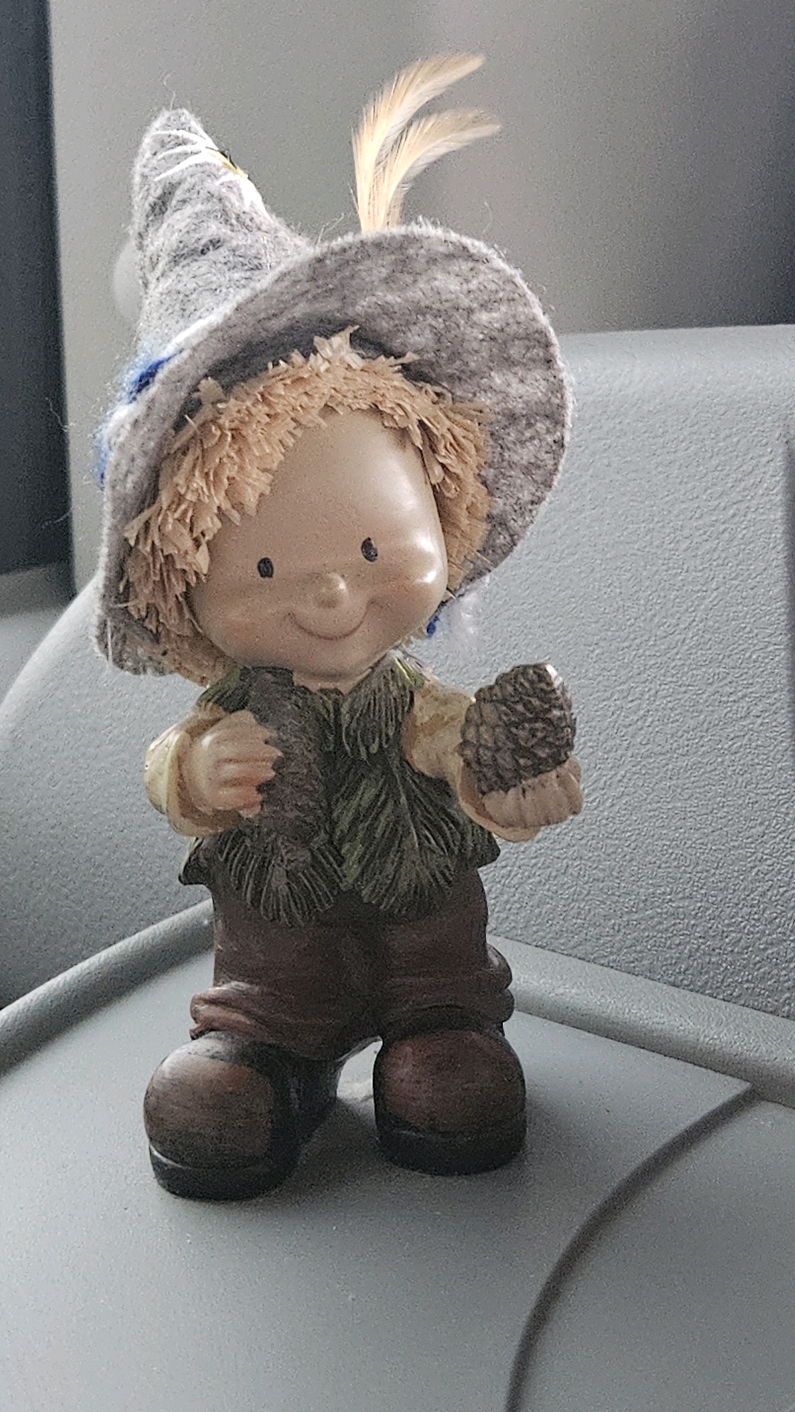 A full-body, eye-level, slightly angled studio shot shows a small, standing gnome figurine on a gray surface. The gnome wears a conical, gray woolen hat adorned with white stitching and two light brown feathers. It has a round, smiling face with black dot eyes and a small nose. The gnome's body is covered by a cream-colored shirt and a dark green vest with leaf-like patterns. Its lower body is clad in brown trousers and large, round, brown boots. In its right hand, the gnome holds a small, brown pinecone. The background is out of focus, showing a textured gray surface on the right and a dark vertical element on the left. The figurine is positioned on a textured gray surface that appears to be a table or shelf. The presence of this figurine does not provide any information about its location in Šarišské Bohdanovce, Slovakia.