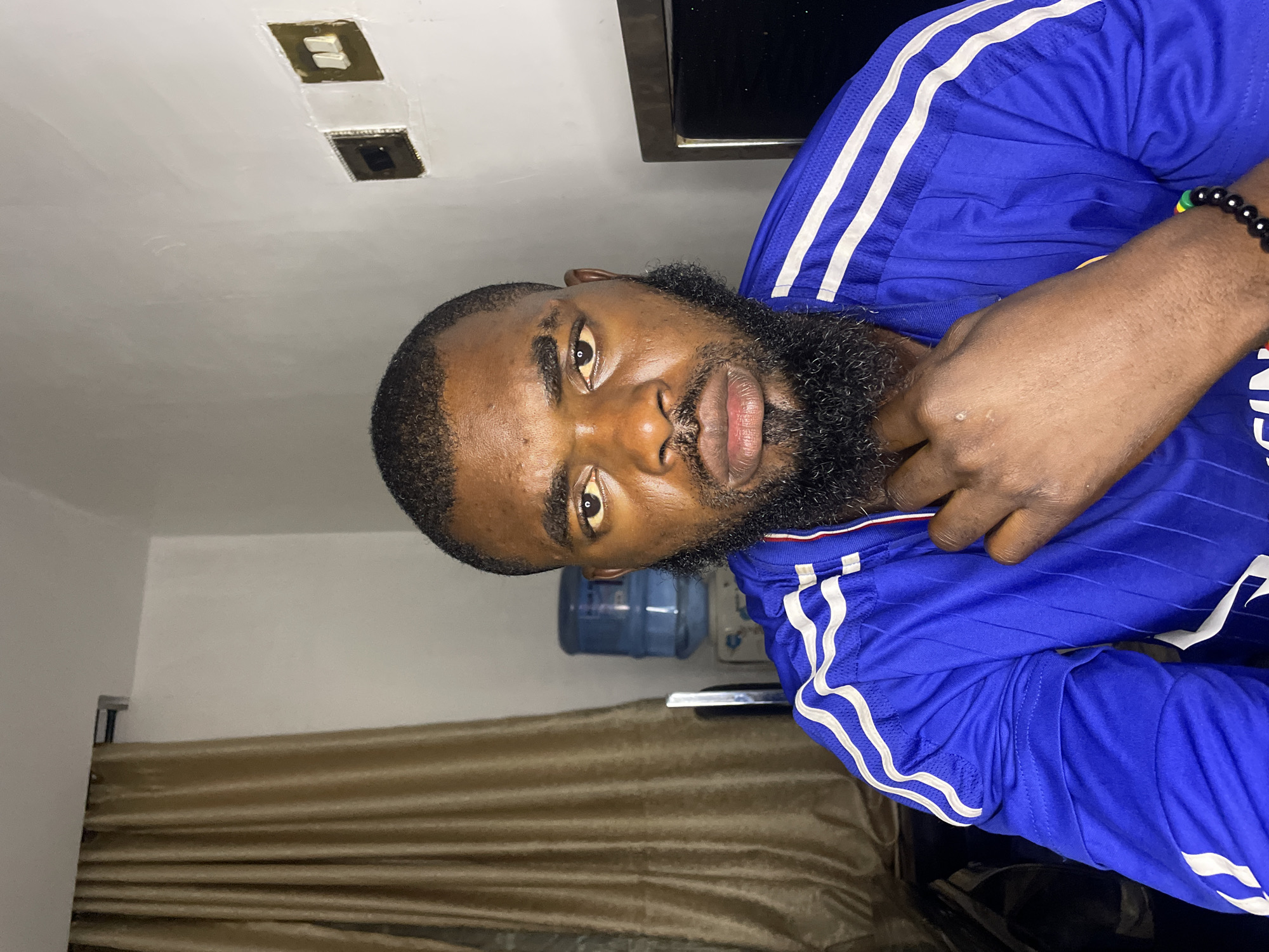 A man with a beard and short hair is seated in front of a light-colored wall and a brown curtain. He is wearing a blue athletic shirt with white stripes on the sleeves. A blue water dispenser is visible behind him to the left. To the right, a dark rectangular object, possibly a television screen, is partially visible. Electrical outlets are mounted on the wall behind the man. The overall lighting suggests an indoor environment, possibly a room in Jos, Nigeria. The man is looking directly at the camera with his hand resting near his chin.