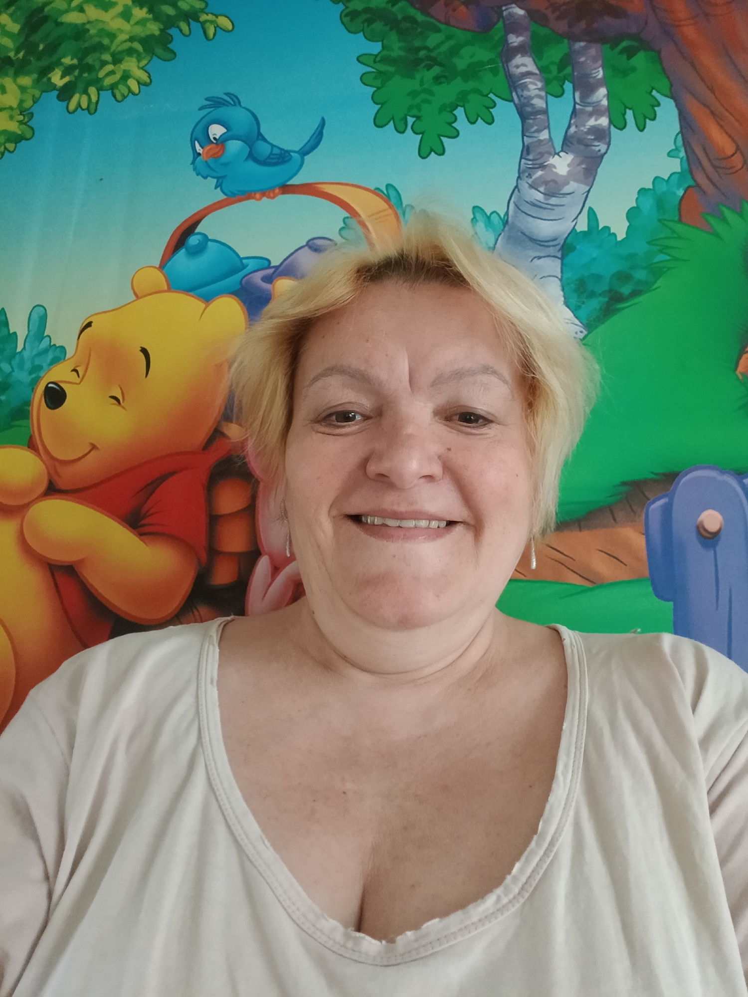A light-skinned woman with short blonde hair is smiling at the camera. She is wearing a light-colored shirt with a low neckline. Behind her is a colorful cartoon backdrop featuring Winnie the Pooh and other cartoon elements like a bluebird, a tree, and other forest scenery. The location and city of Красный Маяк, Russia are not identifiable from the provided image.