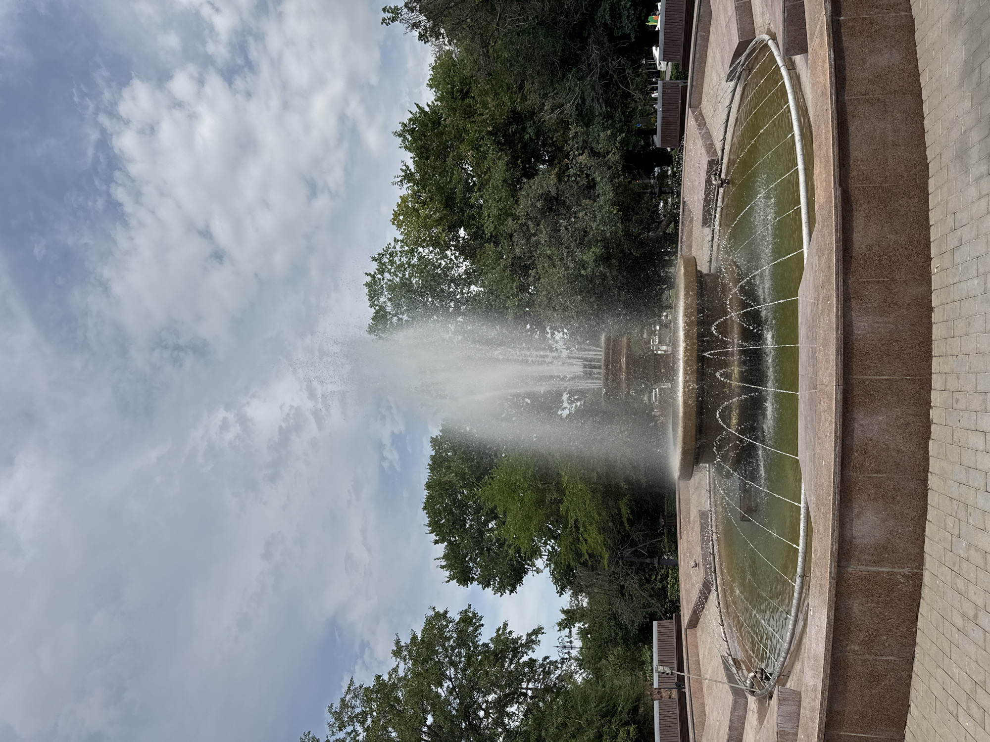 The image shows a large, tiered fountain in an outdoor urban setting. The fountain is made of stone, with a central spray of water reaching high into the air. The lower basin of the fountain is circular and filled with murky green water. Around the fountain, there are paved pathways and several wooden benches. In the background, there are lush green trees and a cloudy sky. The style of the fountain and the presence of benches suggest it is located in a park or public square. The question asks to identify the location as Almaty, Kazakhstan. This information cannot be verified from the image alone.