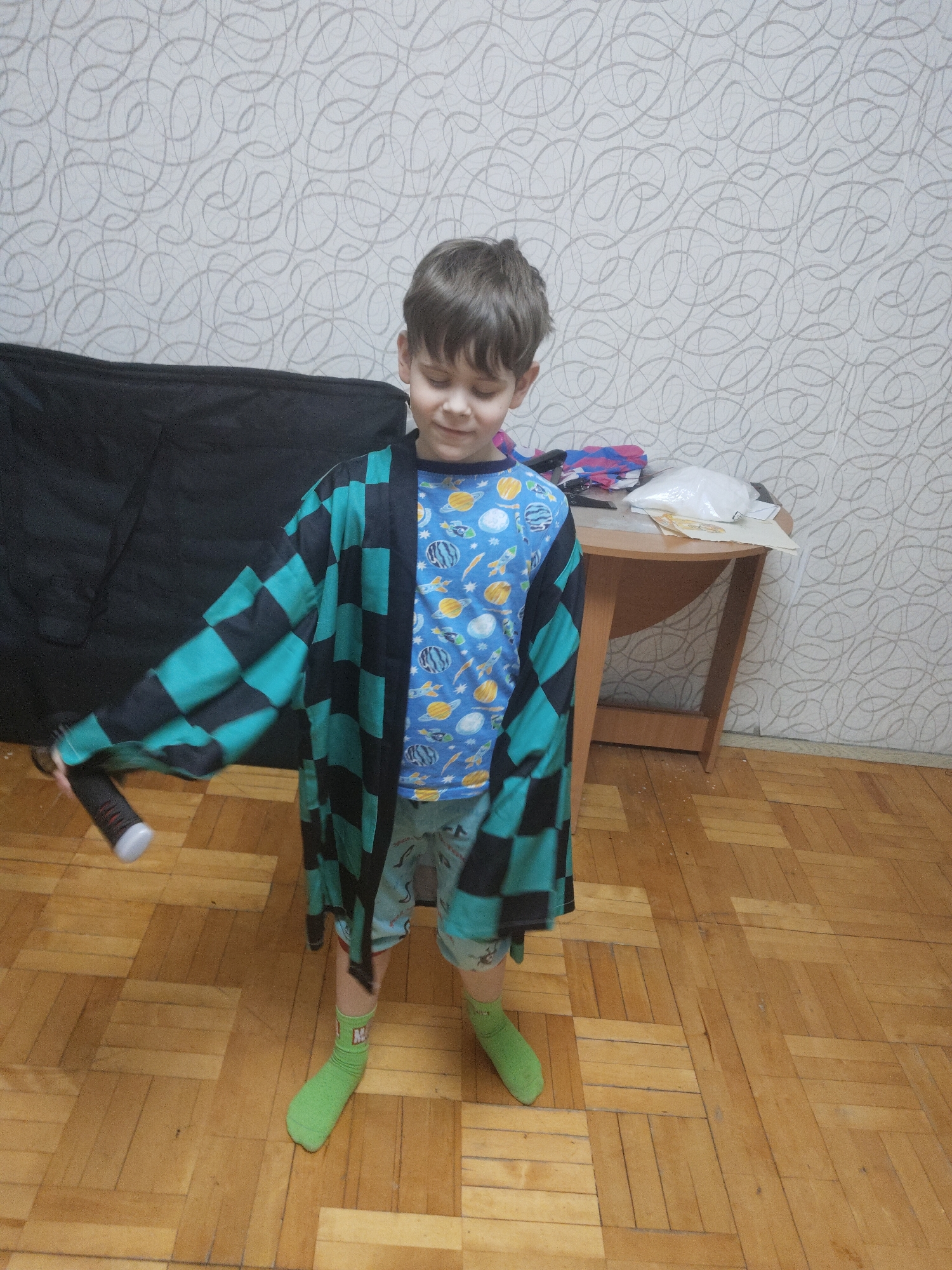 A young boy stands on a wooden floor, wearing a blue shirt with a pattern of planets and spaceships, checkered shorts, and mismatched green socks. He also wears a black and green checkered robe. In his hand, he holds a black object. Behind him, there's a black object, a small brown table with items on it, and a wall with patterned wallpaper. The location is an indoor space with wooden flooring. The city and state are not known.