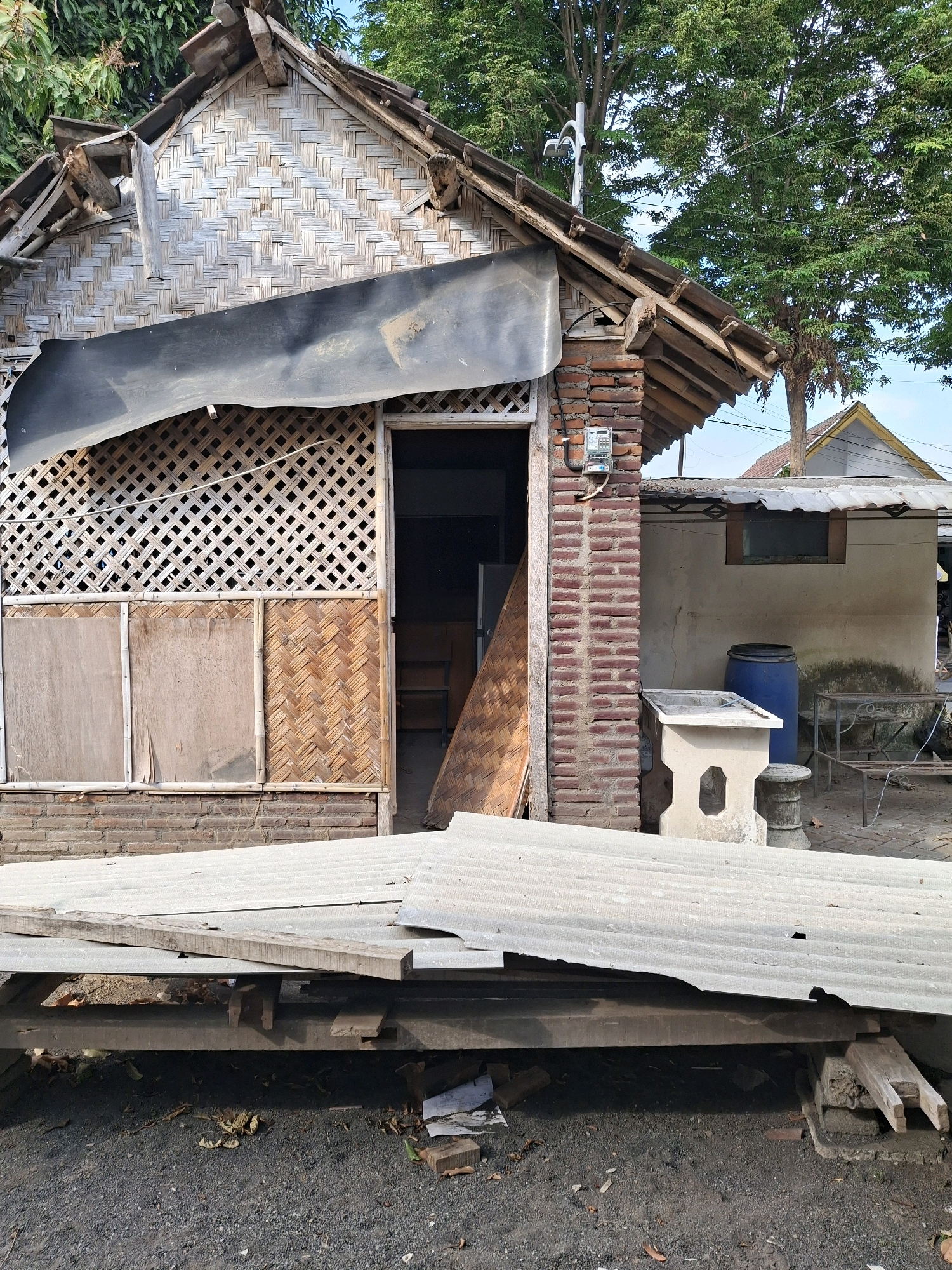 The image depicts a partially constructed or dilapidated building, likely a dwelling, in Lumajang, Indonesia. The structure features walls made of woven bamboo and some brickwork. The roof appears to be a combination of corrugated metal and what looks like traditional woven materials, with some sections damaged or missing. A black tarp is draped over a portion of the wall.
In the foreground, there is a stack of corrugated metal sheets and wooden planks, suggesting ongoing construction or materials for repair. The ground is covered with gravel and scattered debris, including leaves and a piece of wood.
To the right of the main structure, there is another building with a lighter-colored wall and a metal roof. Adjacent to it, a blue plastic barrel, a concrete sink on a pedestal, and a metal table with chairs are visible. An electricity meter is mounted on the brick pillar of the main building. Tall trees with lush green foliage are present in the background, indicating an outdoor setting. The overall scene conveys a sense of rustic simplicity and perhaps a state of disrepair.