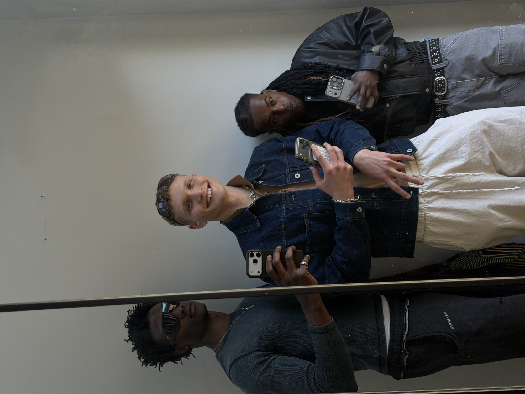 Two individuals are captured in what appears to be a mirror selfie, positioned vertically against a plain, light grey wall. The lower portion of the image shows their reflections in a dark, highly polished surface, possibly a floor or another mirror. The setting suggests an indoor environment, likely in London, United Kingdom.
On the left, a person with fair skin and short, light-colored hair smiles warmly. They are wearing a dark denim jacket over a lighter top, and loose-fitting white pants with visible drawstrings. Sunglasses are perched on top of their head, and they have a silver chain necklace and bracelet. Their right hand holds a smartphone with a distinctive white, circular-textured case, and their left hand forms a peace sign.
To the right, another person with dark skin and long dreadlocks looks directly at the camera with a neutral to slightly serious expression. They are dressed in a black leather jacket, grey cargo-style pants, and a prominent black belt with multiple silver eyelets and a large buckle. This individual also holds a smartphone, which has a solid, light-colored case.
In the reflection below, the first person's denim jacket and white pants are visible. The second person's reflection shows them wearing a dark grey ribbed long-sleeve top and dark joggers. Visible text on the joggers in reflection reads "EMOH AVM" (reversed for "MVA HOME"). Both subjects are equipped with modern smartphones, indicating a contemporary setting. The overall mood is casual and fashion-conscious.