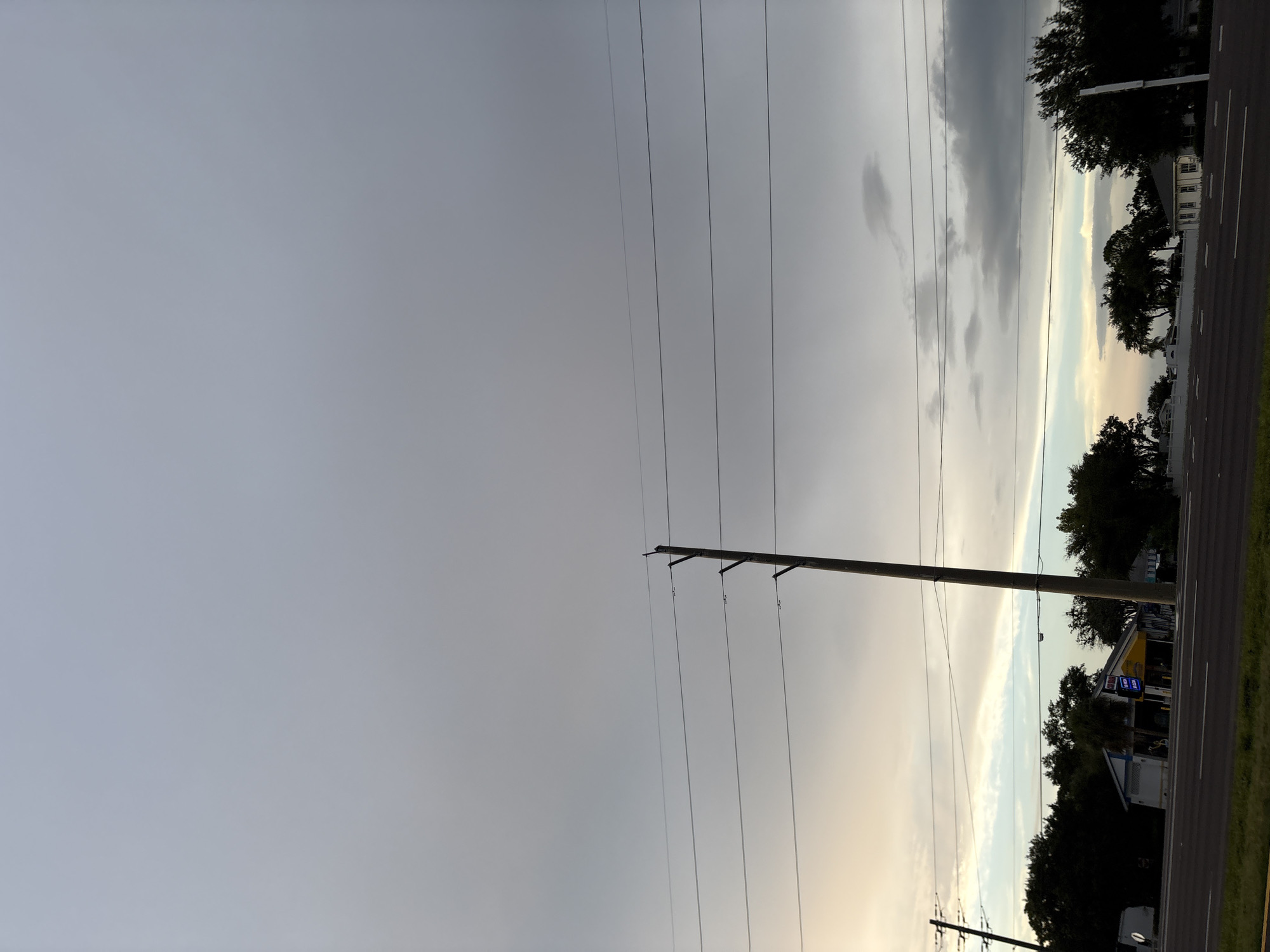 A daytime, full-frame, eye-level shot depicts a vast, overcast sky with a soft gradient of grey and light blue. Several power lines crisscross the frame, suspended from tall wooden utility poles. In the mid-ground, a row of dark green trees lines the horizon, partially obscuring a few buildings. One building, a light blue and yellow structure with a blue sign, is visible on the left. A paved road with white lane markings runs along the bottom of the frame, suggesting it is a street or highway. The scene is static, with no discernible human activity or immediate environmental changes. The location appears to be on a road in Bradenton, United States, possibly near a commercial area due to the presence of buildings.