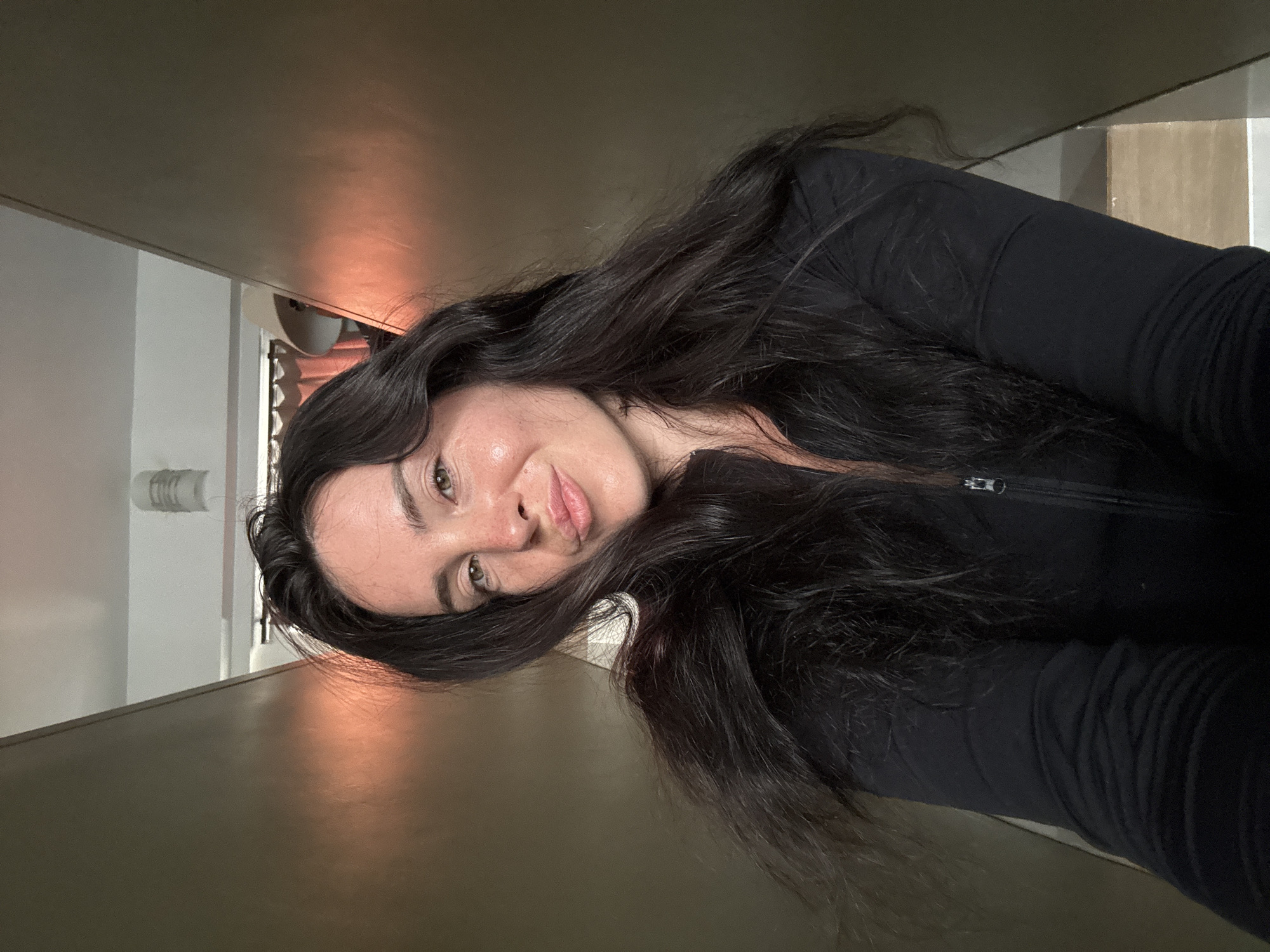 In a selfie taken by a woman with long, dark hair, she is positioned in the center of the frame, her head tilted slightly as she makes a kissing face. Her eyes are open, and she wears a black zip-up jacket. The background suggests an indoor environment, possibly a hallway or between two structures, with warm lighting casting a reddish glow on the walls. No specific elements in the image indicate a location in North Kuta, Indonesia.