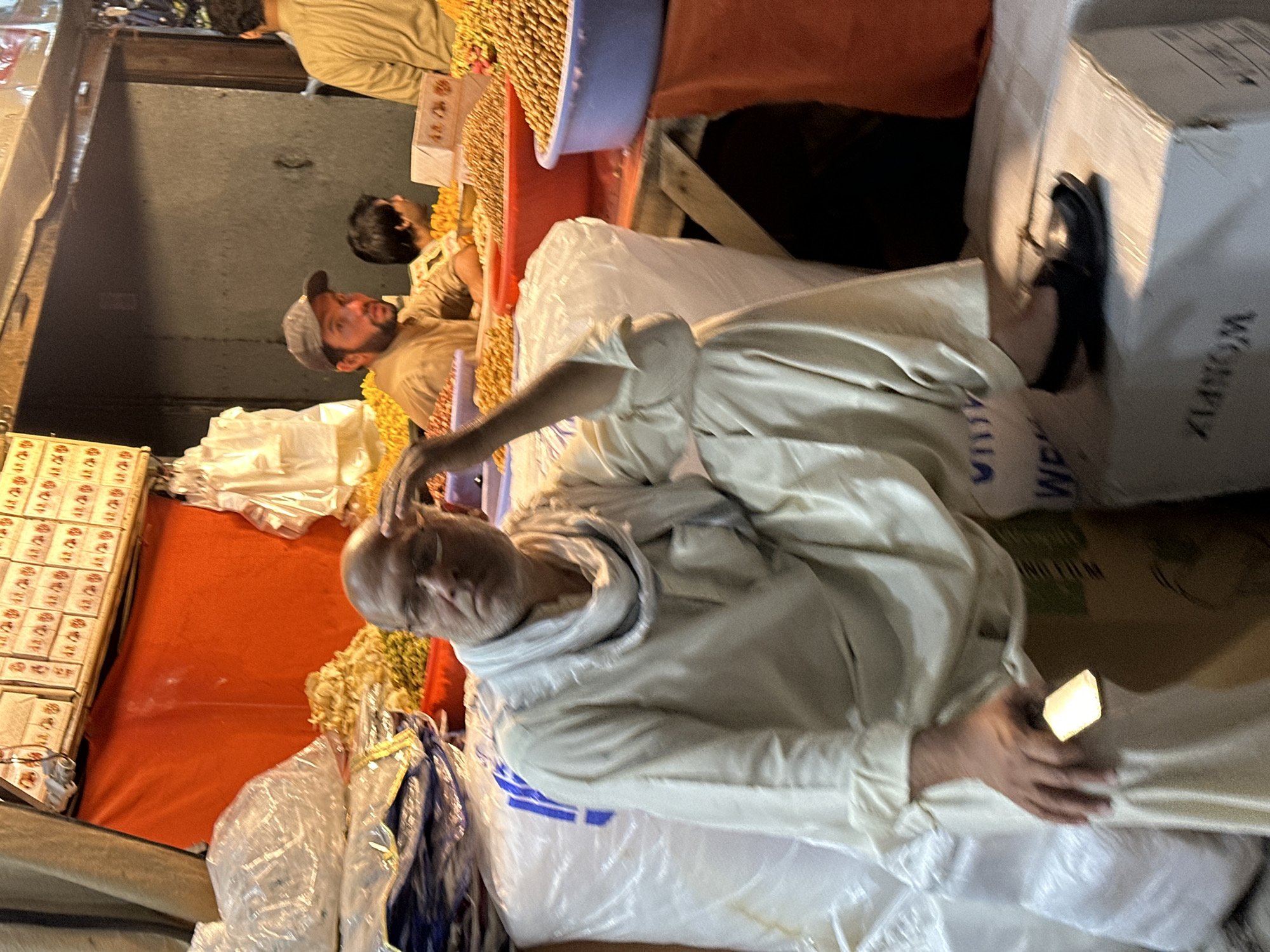 A medium shot of a man in an off-white shalwar kameez seated on a pile of white bags at an outdoor market, presumably in Quetta City Tehsil, Pakistan. He is bald and adjusting his headwear. Other people are visible in the background near what appear to be goods for sale, possibly foodstuffs. A box with "WORKPIX" visible is to the right of the man.