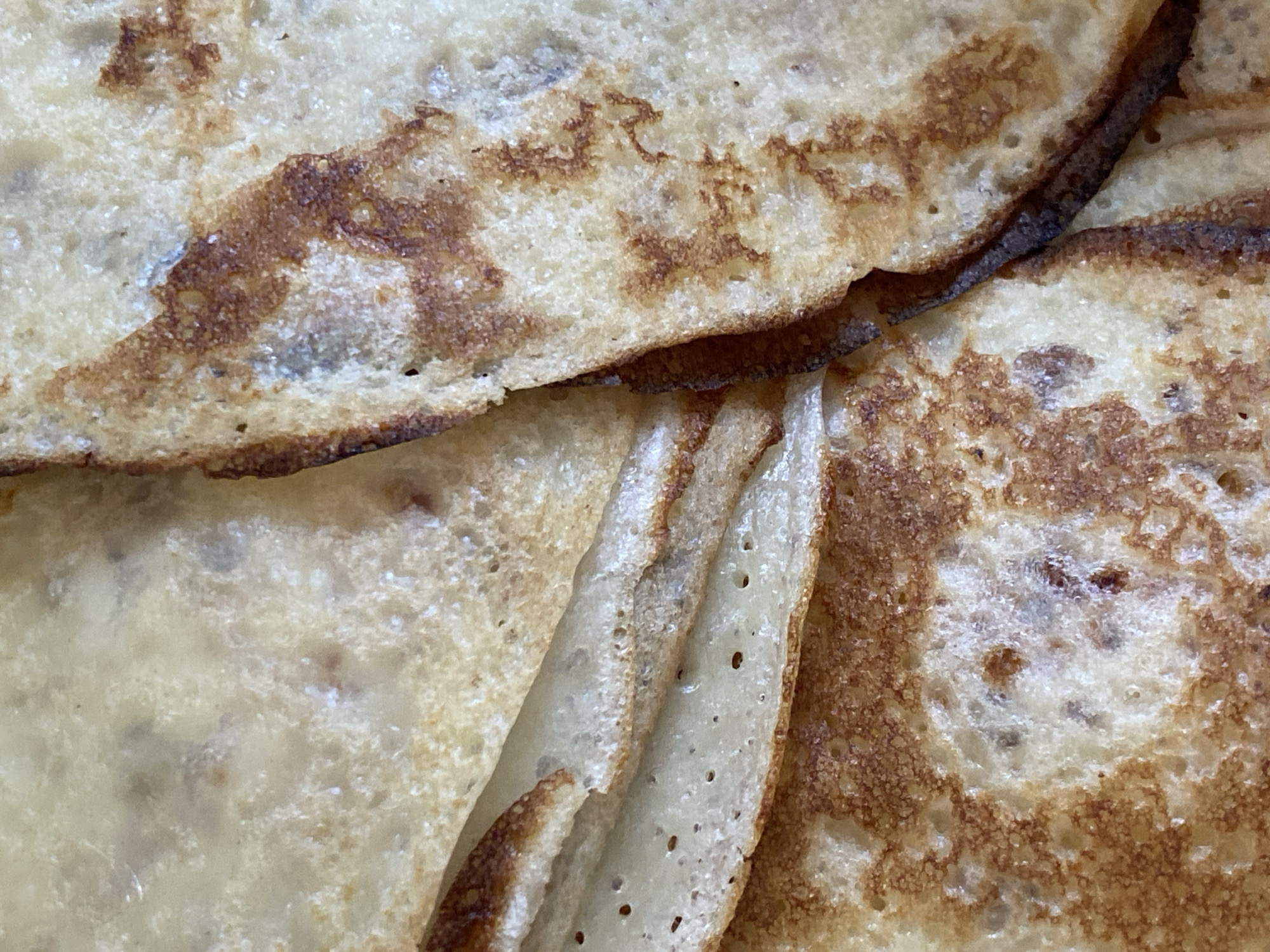 The image displays a detailed, overhead view of several thin, overlapping cooked food items, identified as pancakes or blini, situated within the city of Chaikovsky, Russia. The items exhibit a primary color palette ranging from beige to light golden-brown, punctuated by irregular, darker brown surface caramelization and browning patterns. Visible surface textures include numerous small, circular pores and areas of varying translucency, particularly where the layers fold. The edges of the stacked items show increased browning and a crisper appearance. No human subjects, animals, or other distinct environmental objects are present within the frame.