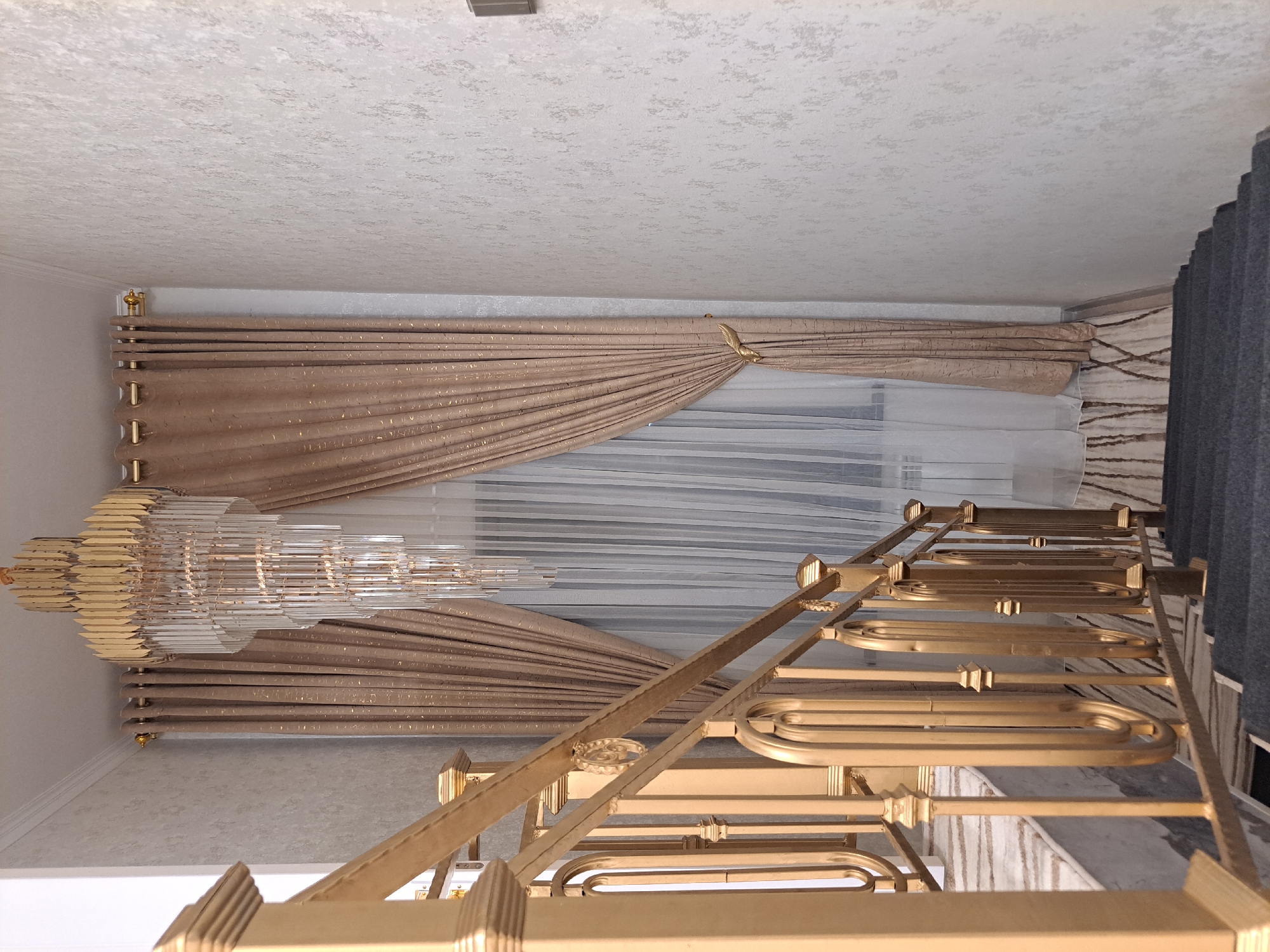 The image depicts a staircase with a golden railing and dark gray carpeted steps. The railing features ornate, rounded metalwork. Above the staircase, a crystal chandelier hangs from the ceiling, casting light on the scene. To the right of the staircase, a window is adorned with heavy, shimmering beige curtains that are pulled to the side, revealing sheer white curtains underneath. The walls are textured with a light, speckled pattern. This interior scene is located in Chirchik, Uzbekistan.