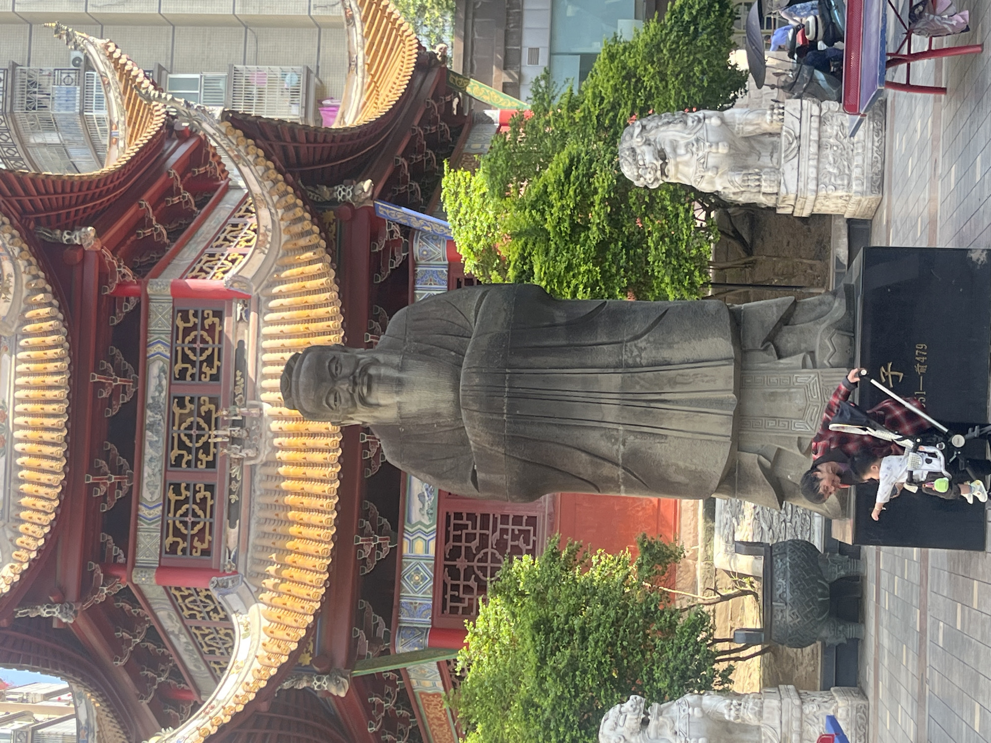 This image captures an outdoor scene featuring a large, stately stone statue, traditional Chinese architecture, and people, set in an urban environment.
The central subject is a tall, dark grey stone statue of an elderly man, likely a revered historical or philosophical figure, depicted in traditional robes and a cap, with a calm and dignified expression. The statue stands on a dark, rectangular base. To its left is an ornate, multi-tiered Chinese pavilion or temple structure, characterized by its sweeping, golden-tiled roofs, rich red pillars, and intricate decorative patterns in blues, whites, and gold on its panels and window lattice work.
Flanking the main statue are smaller, white stone statues of mythical lion-like creatures on pedestals, one clearly visible to the right and another partially seen in the lower left. Lush green bushes and trees are interspersed around the statues and architecture, providing natural contrast.
In the foreground, near the base of the main statue, a person wearing a red and black plaid shirt is pushing a baby stroller with a child inside, suggesting a casual visit or family outing. A black umbrella, a red metal stand, and a dark, ornate urn-like object are also visible in this area. The ground is paved with light grey patterned tiles.
In the background, modern multi-story buildings with numerous windows rise above the traditional structures, indicating a blend of historical and contemporary elements within an urban setting. The scene is brightly lit by daylight, suggesting a clear, sunny day.
Visible Chinese text on the black base of the main statue includes the character "子" (zǐ), likely part of a name such as "孔子" (Kǒngzǐ, Confucius), and below it, "一首479". Given the context of Jiangjin, China, this scene likely depicts a historical or cultural site dedicated to a prominent figure.