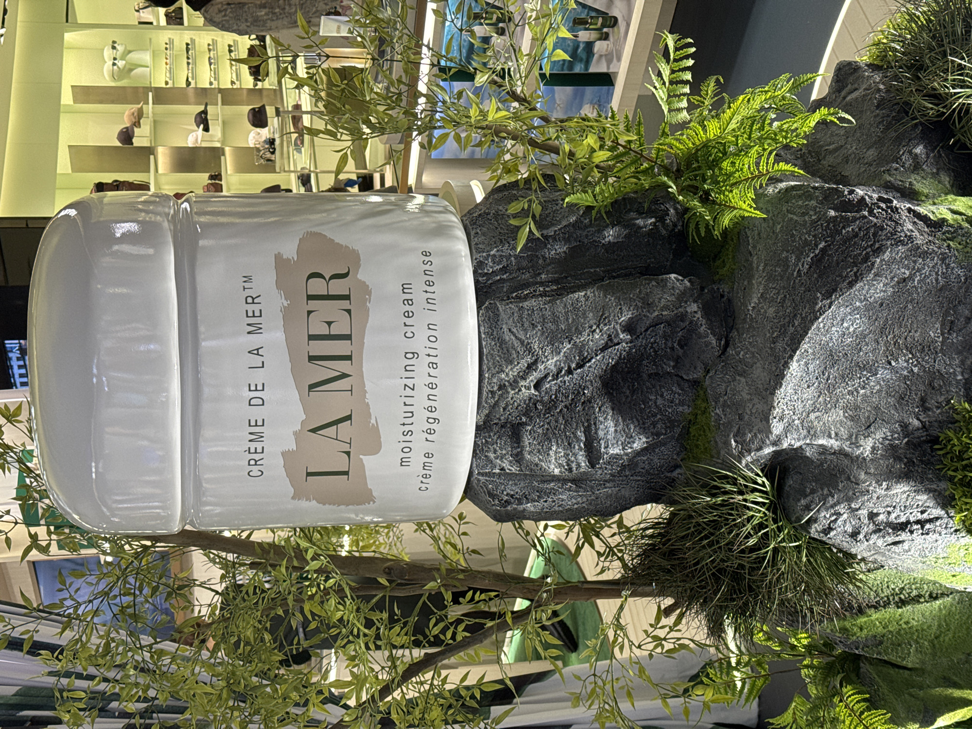 A close-up shot features a white jar of La Mer moisturizing cream prominently displayed on a rocky, mossy surface, surrounded by artificial greenery. The jar has "LA MER" in large, tan letters across its front, with "CRÈME DE LA MER™" to the left and "moisturizing cream crème régénération intense" to the right. Behind the cream jar, shelves are visible with various products, suggesting a retail or display setting. The scene is lit, creating shadows and highlights on the rocks and plants. The overall aesthetic evokes a luxurious, natural, and spa-like atmosphere, possibly within a high-end retail store or a beauty exhibition in Nong Prue Subdistrict, Thailand. The image is oriented sideways.