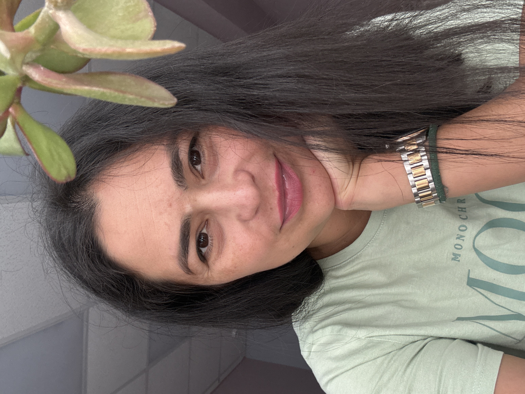 A woman with long, dark hair and brown eyes is smiling and looking at the camera. She is wearing a light green t-shirt with the word "MONOCHR" visible. A gold and silver watch is on her left wrist, which is placed under her chin. A succulent plant is visible in the upper left corner of the image. The background is light grey and blurry. The location is not indicated within the image. It is not possible to confirm its location in Chaikovsky, Russia, from the information given.