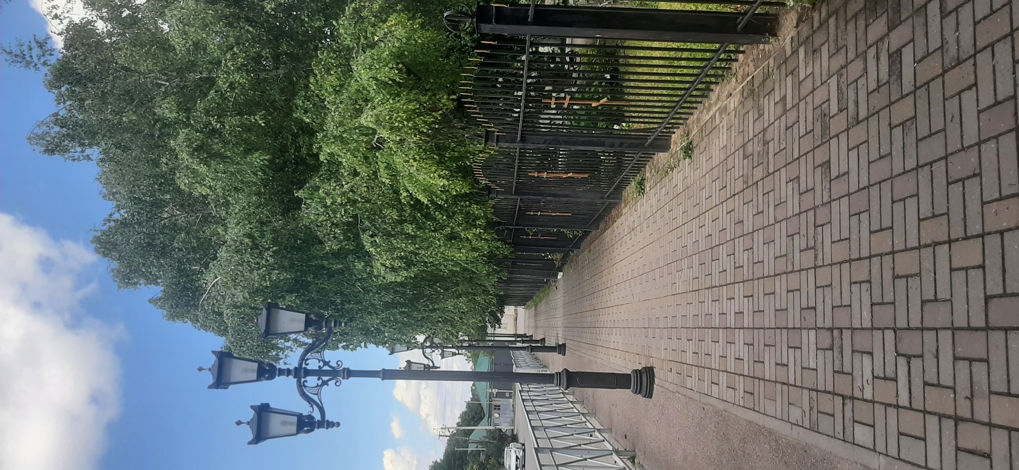 The image shows a paved sidewalk with a row of decorative lampposts on the left and a black metal fence with ornate tops and crosses on the right. A large tree with dense green foliage is visible behind the fence. The sky is blue with scattered white clouds. In the distance, a white car is driving on a road next to the sidewalk, and some buildings with green roofs are visible. The sidewalk is made of rectangular bricks laid in a herringbone pattern. The overall scene depicts an outdoor urban environment on a bright day.