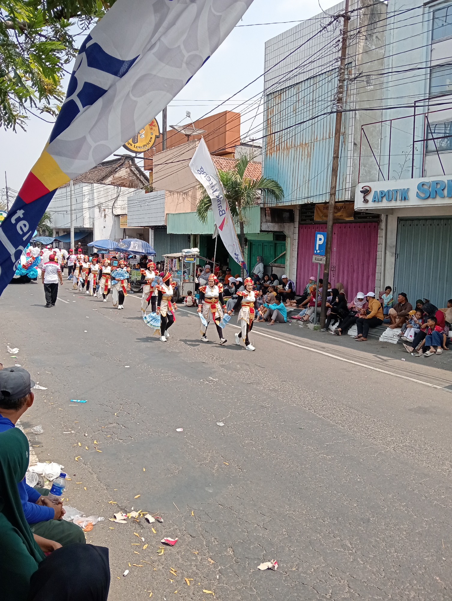 The image depicts a street scene in Demak, Indonesia, likely during a parade or festival. A group of performers in ornate, gold and white costumes with red accents are marching down the street. They are dressed in helmets and some are holding fans. In the background, spectators are seated along the side of the road, some under umbrellas. Buildings line the street, with signs indicating businesses such as "APOTIK SRI." A large banner with blue and white patterns hangs in the foreground, partially obscuring the view of the procession. The street is paved and shows signs of wear. The weather appears to be sunny.