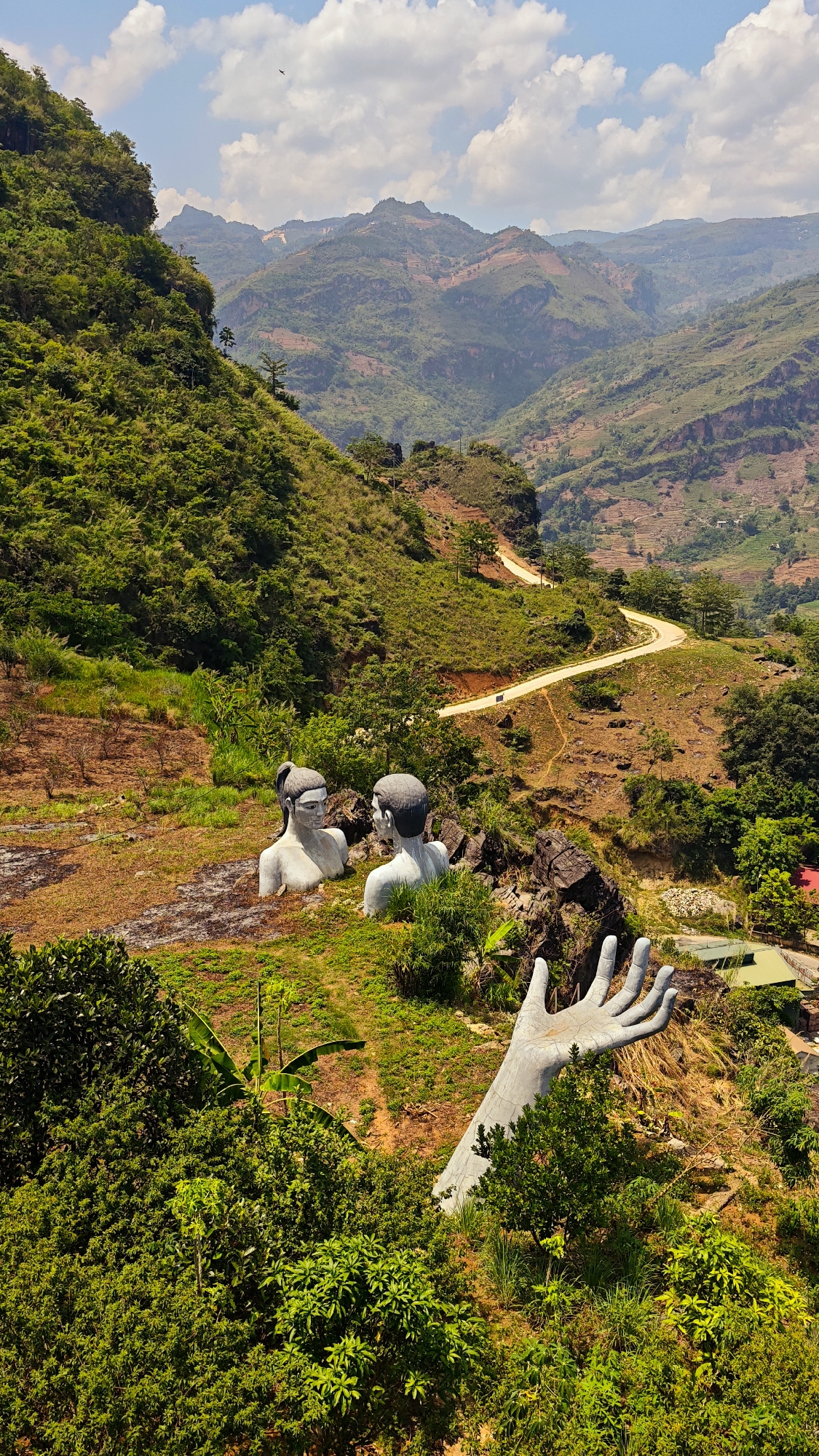 The image shows a landscape in Cốc Pài, Vietnam. Two bust sculptures of a man and woman face each other. A large sculpture of a hand reaches out from behind dense vegetation. A winding road traverses the hillside in the background, leading into a valley of forested mountains under a partly cloudy sky.