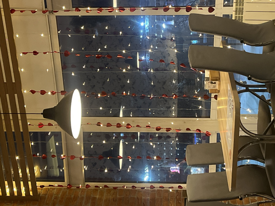 This image captures the interior of a dining establishment in Chaikovsky, Russia, looking outwards through a large window during nighttime. The window displays extensive festive decoration, featuring numerous vertical strings of small, warm white fairy lights interlaced with red heart-shaped ornaments. A single, illuminated dark grey industrial-style pendant lamp hangs centrally from the ceiling, positioned in front of the window.
To the left, a portion of a brick wall is visible. Above the window, wooden horizontal blinds or slats are present. In the foreground, a rectangular table with a light-colored wooden top and a silver metal base is situated. Four chairs with grey upholstered backs and silver metal frames are arranged around this table, two on each side, all currently vacant. On the table, a wooden caddy and several paper items are present. A white radiator is visible at the bottom right corner of the frame.
Through the decorated window, the exterior scene is dark, illuminated by numerous distant artificial lights from buildings and streetlights, which cast reflections on the glass. Snow is evident on the ground outside. No individuals or active interactions are depicted within the frame.