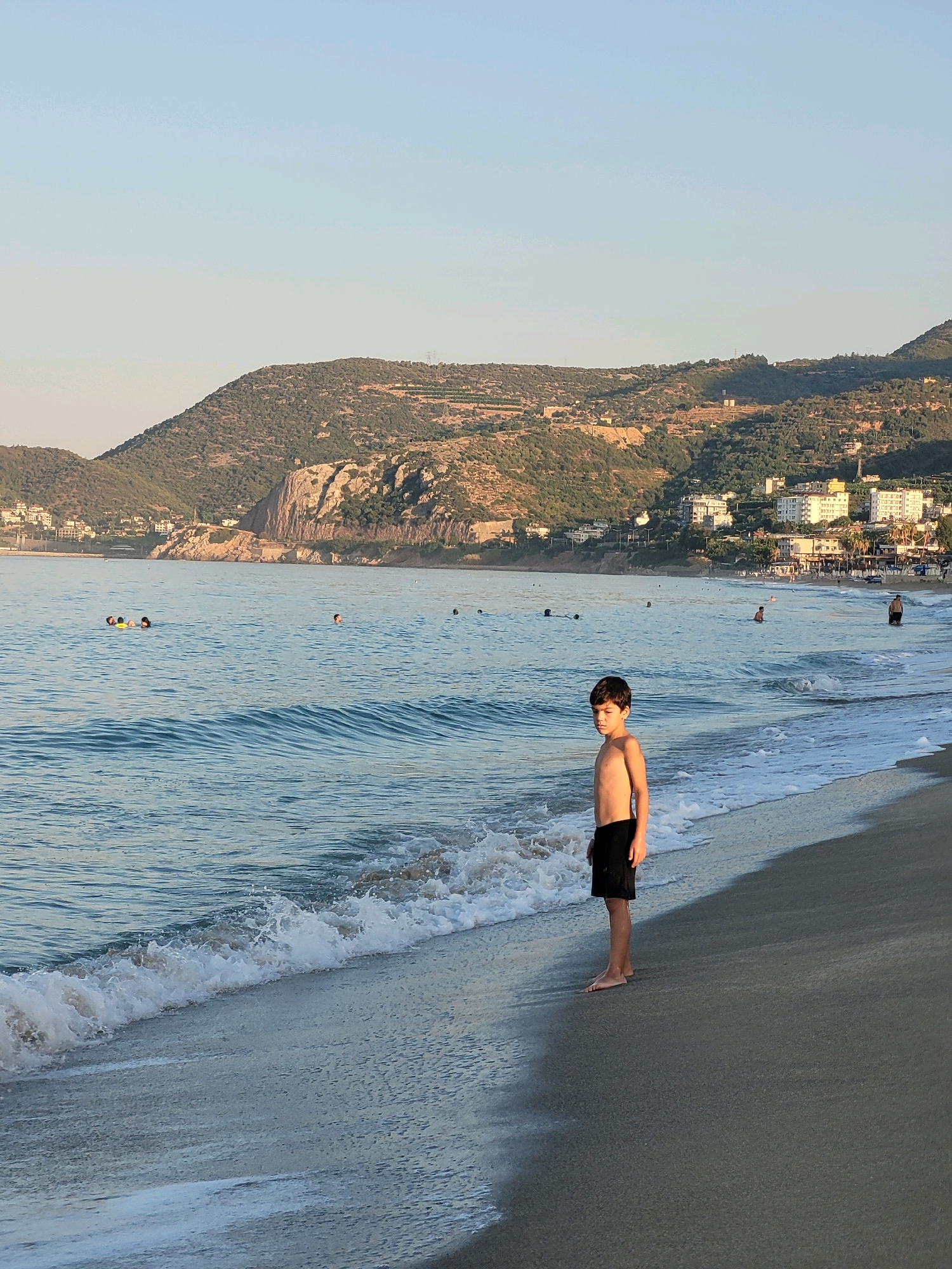 A young boy stands on a sandy beach in Alanya, Turkey, looking towards the viewer. He is wearing black swim trunks. The ocean is to his left, with gentle waves lapping at the shore. In the distance, several people are swimming or wading in the water. Across the sea, a coastline of hills is visible, dotted with buildings and vegetation. The sky above is a clear, pale blue. The overall scene suggests a relaxed, late afternoon or early morning at a coastal resort.