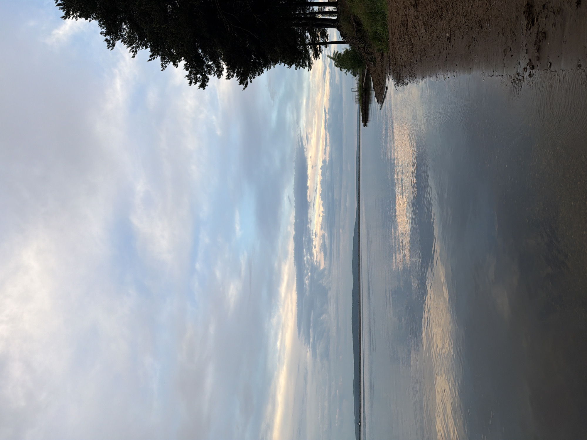 The image depicts a body of water, likely the Kama River, adjacent to a sandy shore and a treeline. The sky is filled with clouds, partially reflecting in the water. A dock or pier extends into the water. The image appears to be taken in the city of Chaikovsky, Russia, based on the location provided. There is no evidence of any people, interactions or actions.