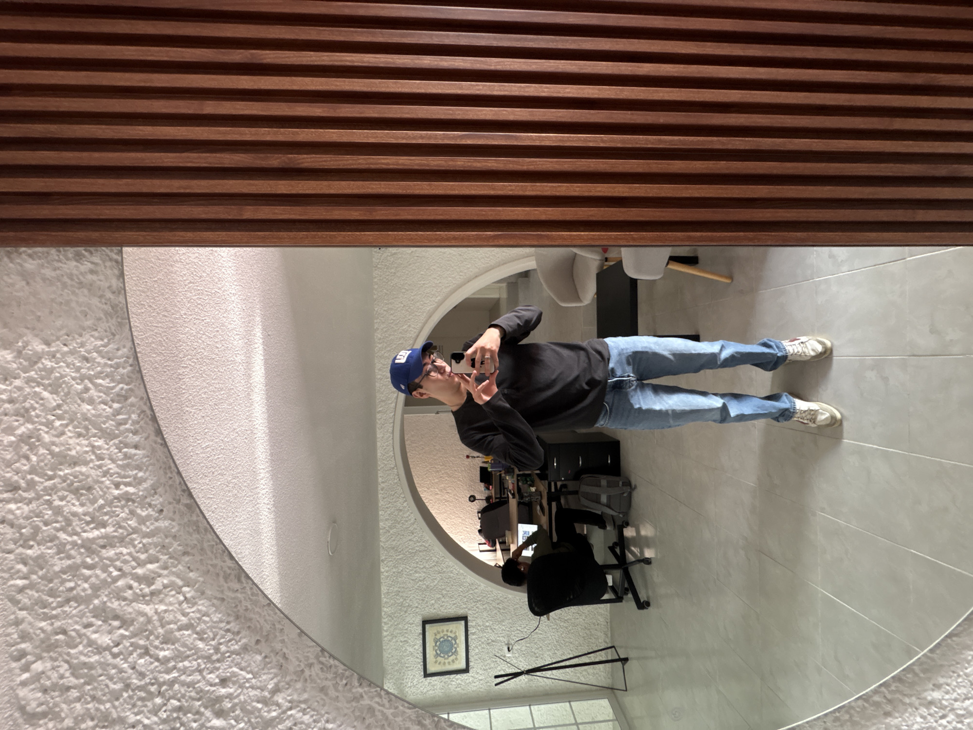 A person is seen in a large, partially circular mirror, taking a selfie with a white smartphone. The image is oriented with the wooden slatted wall panel at the top. The person in the mirror is standing, wearing a blue baseball cap, glasses, a dark long-sleeved top, light blue jeans, and white sneakers. Their expression appears neutral, focused on the phone.
The mirror reflects a modern indoor space, likely an office or communal area. Above the mirror, a prominent wooden slatted wall panel runs horizontally. To the left of the mirror, a textured white wall is visible. Inside the reflection, behind the person taking the selfie, another individual is seated at a desk, facing a computer monitor, suggesting they are working. This desk area includes a black office chair, a dark cabinet or storage unit, and a framed piece of art on the wall below the desk. A light stand or tripod is also visible on the tiled floor. The floor is covered in light-colored square tiles. Faintly visible in the upper part of the reflection are white chairs and a table. The lighting suggests it is daytime, with interior light sources illuminating the room. No discernible text is visible in the image. The setting is a contemporary interior, located in Puebla, Mexico.