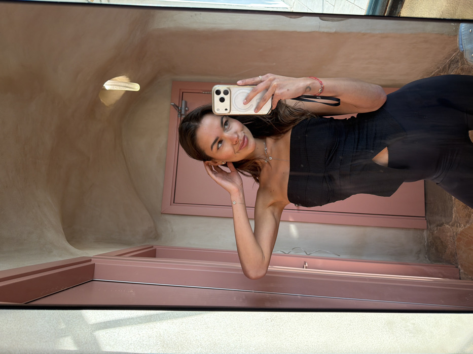 A female subject, appearing in her 20s or early 30s, is captured in a waist-up mirror selfie within an indoor setting. She has light brown, wavy hair and displays a soft smile, gazing towards the reflected camera. Her attire consists of a black strapless top with ruched detailing on the bust and a split front hem, paired with black form-fitting trousers or leggings. She wears stud earrings, a delicate necklace with a small pendant, a bracelet on her left wrist, a thin ring on her right ring finger, and a red string bracelet on her right wrist. Her right hand holds a light-colored smartphone, likely an iPhone with a cream-colored MagSafe case, positioned to take the photograph. Her left hand is raised, with fingers touching the side of her head near her ear.
The background, visible in the mirror's reflection, features a distinctive interior space. The walls and ceiling are textured, appearing in a warm, light beige or tan color, characterized by smooth, curvilinear, and organic architectural forms, reminiscent of a grotto or shell interior. An elongated, vertical light fixture is embedded within the rounded ceiling structure. Two doorways are present, both framed and painted in a distinct dusty rose or muted pink hue. The left doorway appears to be a window or glass door allowing natural light to enter, casting a shadow pattern. The right doorway is a solid pink door equipped with a gold-toned latch mechanism. This specific architectural style, including the organic forms and characteristic pink accents, identifies the location as the Dr. Meller House (also known as the Shell House) in Jaffa, Tel-Aviv-Yafo, Israel.