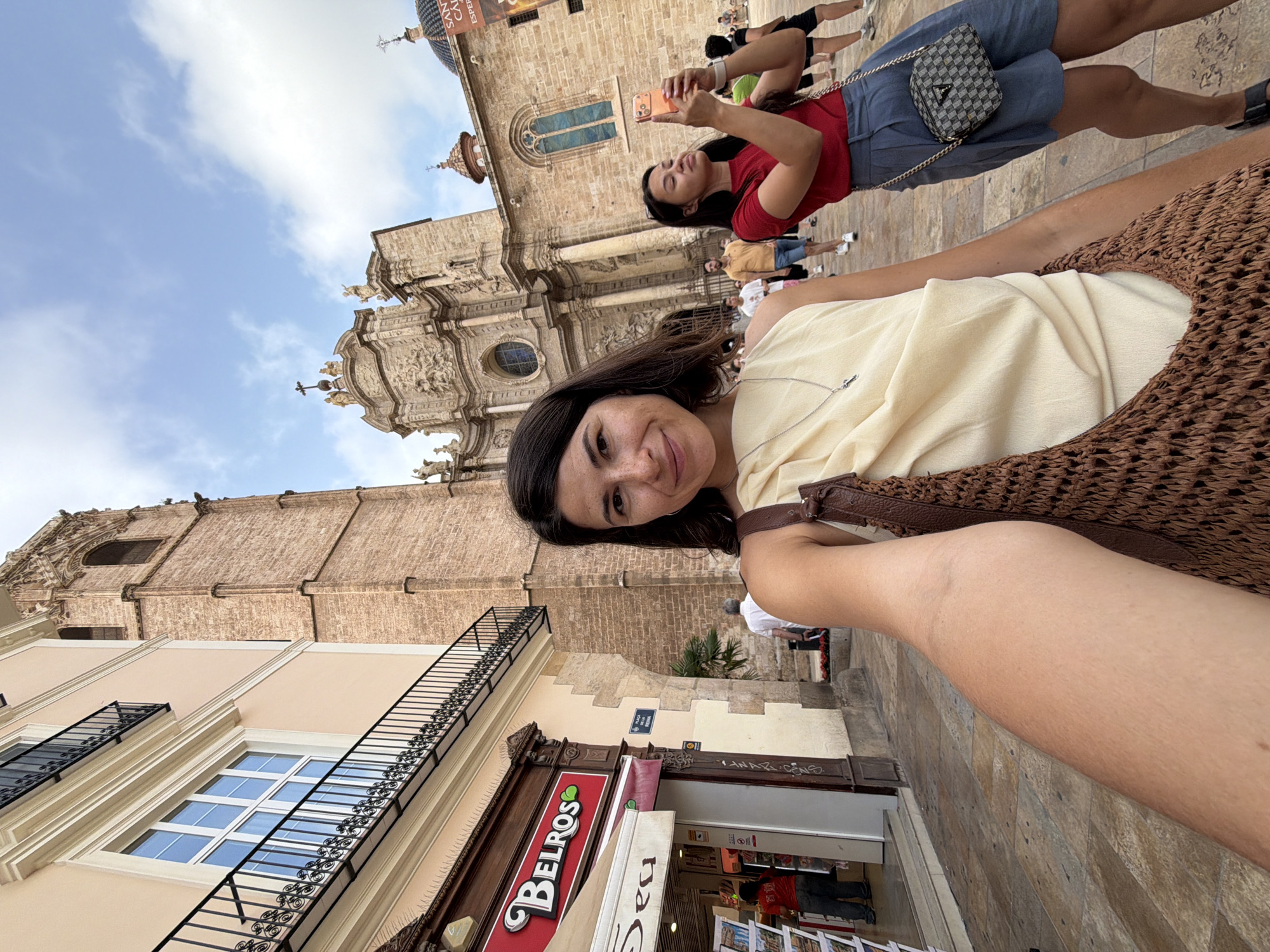 A woman in a light yellow top takes a selfie in an urban plaza with a large cathedral in the background. To her left is a store with a red sign that says "BELROS." Behind her, another woman in a red shirt and blue shorts is taking a photo with her phone. Other people are walking in the plaza. The ground is paved with stone tiles, and the sky is blue with some clouds.