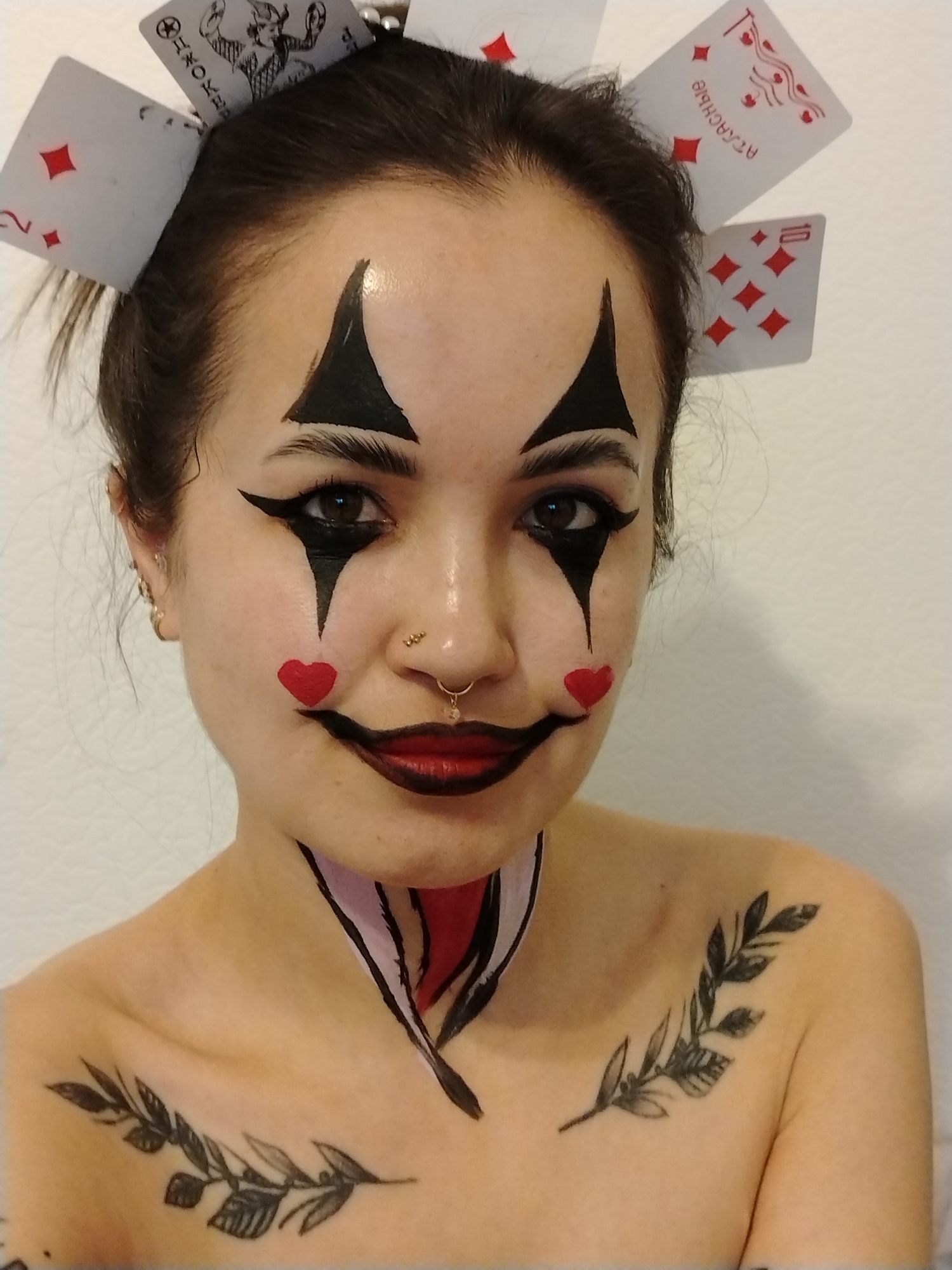A young woman with dark hair pulled back is centered in the frame, her face adorned with clown-like makeup. Black diamond shapes are painted above her eyebrows, with black lines extending down from the corners of her eyes. Small red hearts are painted on her cheeks. Her lips are painted with a dark, exaggerated smile. She has multiple piercings in her nose and ears. Playing cards are positioned around her head, appearing to be attached to her hair. Her collarbone is decorated with black painted patterns, including a branch-like design and geometric shapes. The background is a plain, light-colored wall. There is no indication of the location being Noviy, Russia.