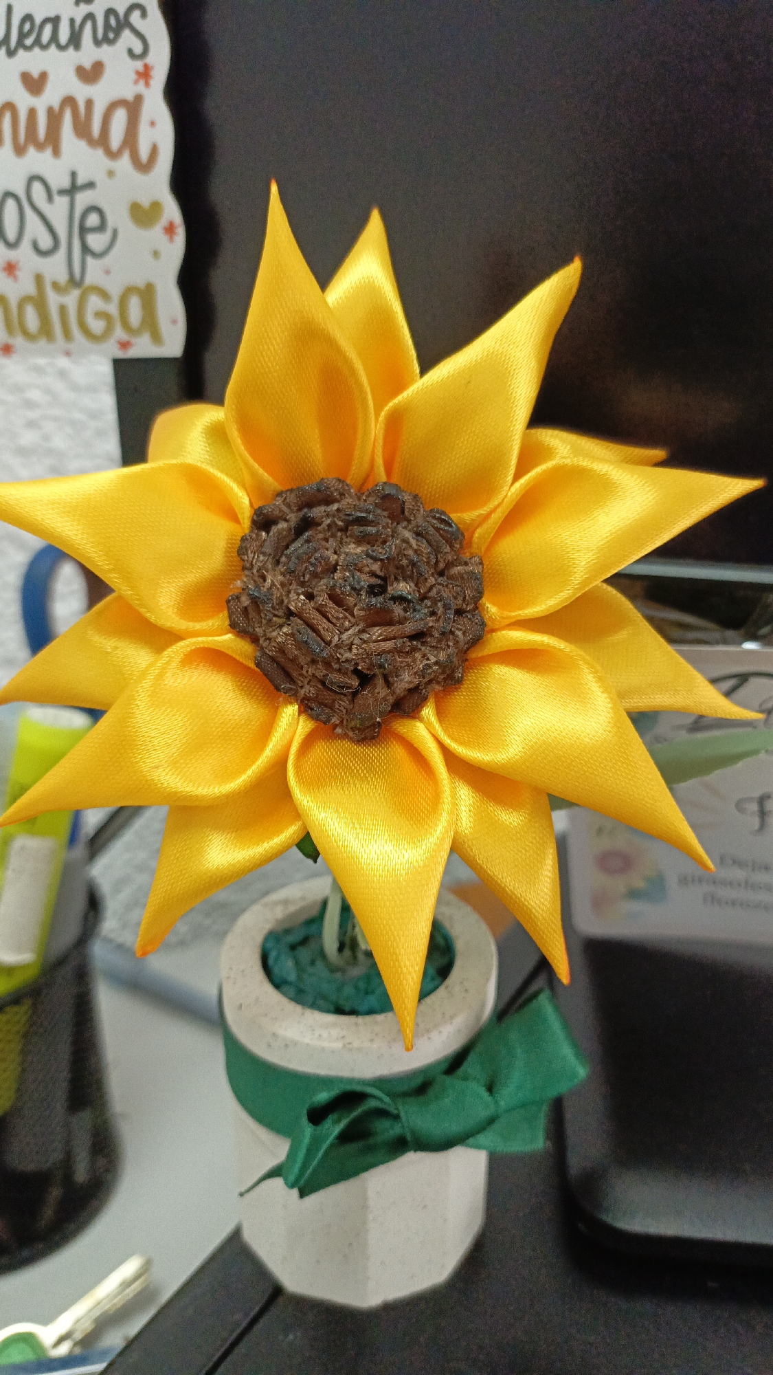 The image shows a crafted sunflower made of yellow ribbon with a brown center, placed in a small, white, geometric-shaped pot. A green ribbon is tied around the pot. The sunflower is positioned on a desk or countertop, with office supplies and a sticker visible in the background. The image was likely taken indoors. I cannot determine the location within Guayana City, Venezuela from the given information.