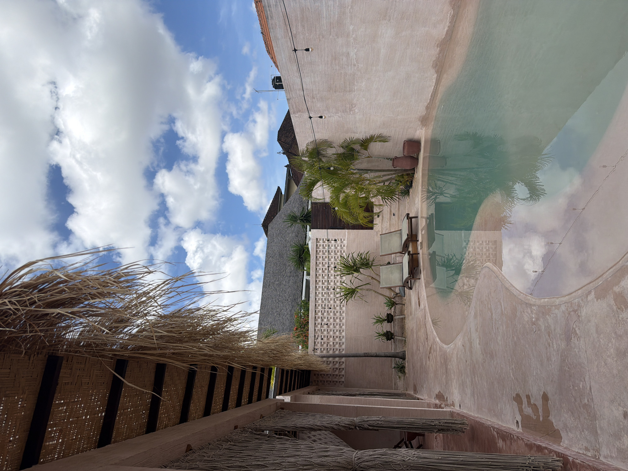 The image shows a poolside area in North Kuta, Indonesia. The pool's irregular shape mirrors the cloudy sky. The area features lounge chairs, potted plants, and palm trees. A woven thatched roof provides shade. The building surfaces have a textured, stucco-like appearance.