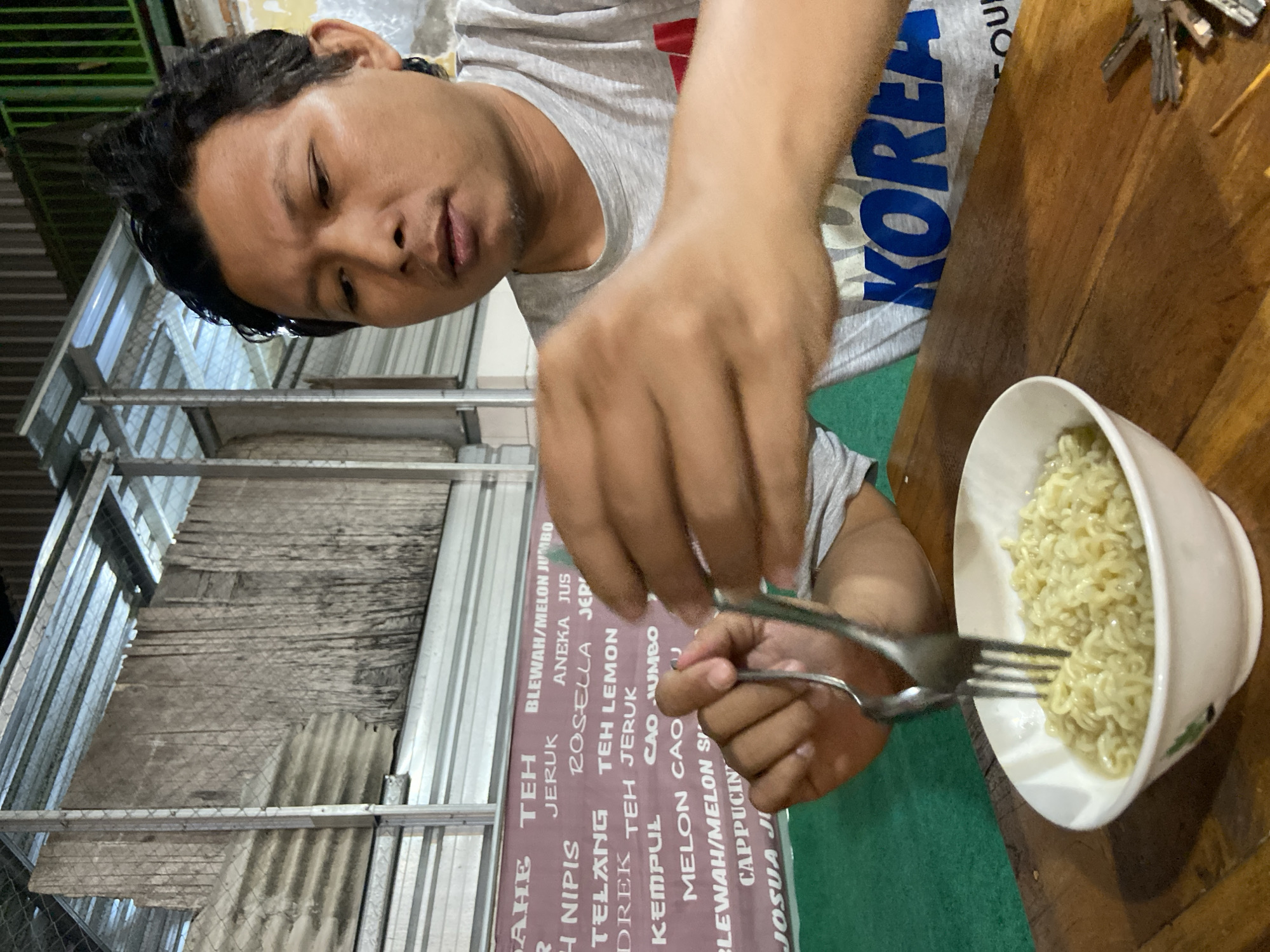 The image shows a person eating instant noodles at what appears to be an eatery in Sidoarjo, Indonesia. The subject, an individual with dark hair, is seen from a high-angle perspective, wearing a grey t-shirt with "KOREA" printed in blue on the chest. They are actively using a fork and spoon held in their right hand to eat instant noodles from a white bowl positioned on a rustic wooden table.
The noodles in the bowl are light yellow and curly, typical of instant varieties. Keys are also visible on the wooden table near the bowl. In the background, a large vertical menu board displays a variety of drinks and food items, with visible text including "BLEWAH/MELON JUMBO," "ANEKA JUS," "ROSELLA JERUK," "TEH LEMON," "MELON CAO," "CAPPUCIN," and "JOSUA." Other listed items are "AHE TEH," "JERUK," "NIPIS," "TELANG TEH LEMON," "DREK TEH JERUK," and "KEMPUL CAO JUMBO." Behind the menu and to the far left, there is an industrial-looking structure made of metal beams and corrugated sheets, suggesting an outdoor or semi-outdoor setting. The lighting suggests it is evening or night, with artificial light illuminating the scene. The person's expression is neutral and focused on their meal.