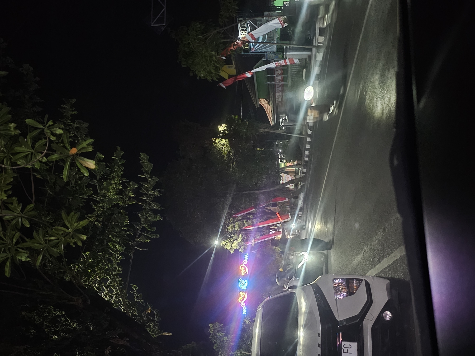 The image captures a nighttime scene on a street in Lamongan, Indonesia, likely near the Alun-Alun, given the festive decorations. The foreground features the interior of a car, with the dashboard and windshield visible. Beyond the car, a white SUV is parked on the left side of the road, its headlights illuminated. A person in dark clothing is standing on the road, facing away from the camera, possibly directing traffic or participating in an event. Further down the road, a motorcycle with its headlight on is approaching.
The street is decorated with vibrant, colorful lights strung across the road, creating a festive atmosphere. Red and white Indonesian flags are prominently displayed on poles along the sidewalk and on buildings in the background. The buildings themselves appear to be low-rise structures, with a tiled roof visible on one of them. Trees line the street, their leaves illuminated by the artificial lights. The road surface is wet, reflecting the lights and creating a glossy effect. The overall impression is of a lively evening, possibly during a celebration or event in the city.