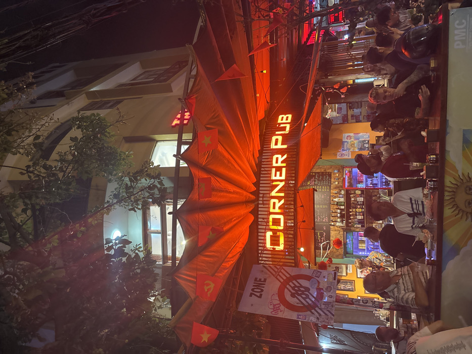 The image depicts a nighttime scene at the "Corner Pub" located in Ngũ Hành Sơn Ward, Vietnam. The pub's facade is dominated by a prominent orange neon sign displaying "CORNER PUB." An expansive red fabric canopy, illuminated from below, extends over the outdoor seating area, adorned with numerous small Vietnamese national flags (red with a yellow star) and several flags of the Communist Party of Vietnam (red with a yellow hammer and sickle).
Beneath the canopy, approximately 15-20 individuals, appearing to be adults of various ages and genders, are visible. Most are seated or standing around a long bar counter in the foreground, engaging in social interaction. Several men are seated at the counter with drinks. A black motorcycle helmet rests on the counter in the lower right. Behind the counter, a well-stocked bar with numerous bottles is brightly lit, and at least one staff member is present.
To the left of the pub's entrance, a large white banner with a "ZONE 38" logo is displayed. Small red lanterns are suspended near the bar. The pub occupies the ground floor of a multi-story building, with dimly lit upper floors visible above the canopy. Dark tree branches and foliage partially obscure the upper portion of the building and night sky. The overall lighting is a vibrant red-orange, emanating from the pub's signage and canopy, contrasting with the darker background.