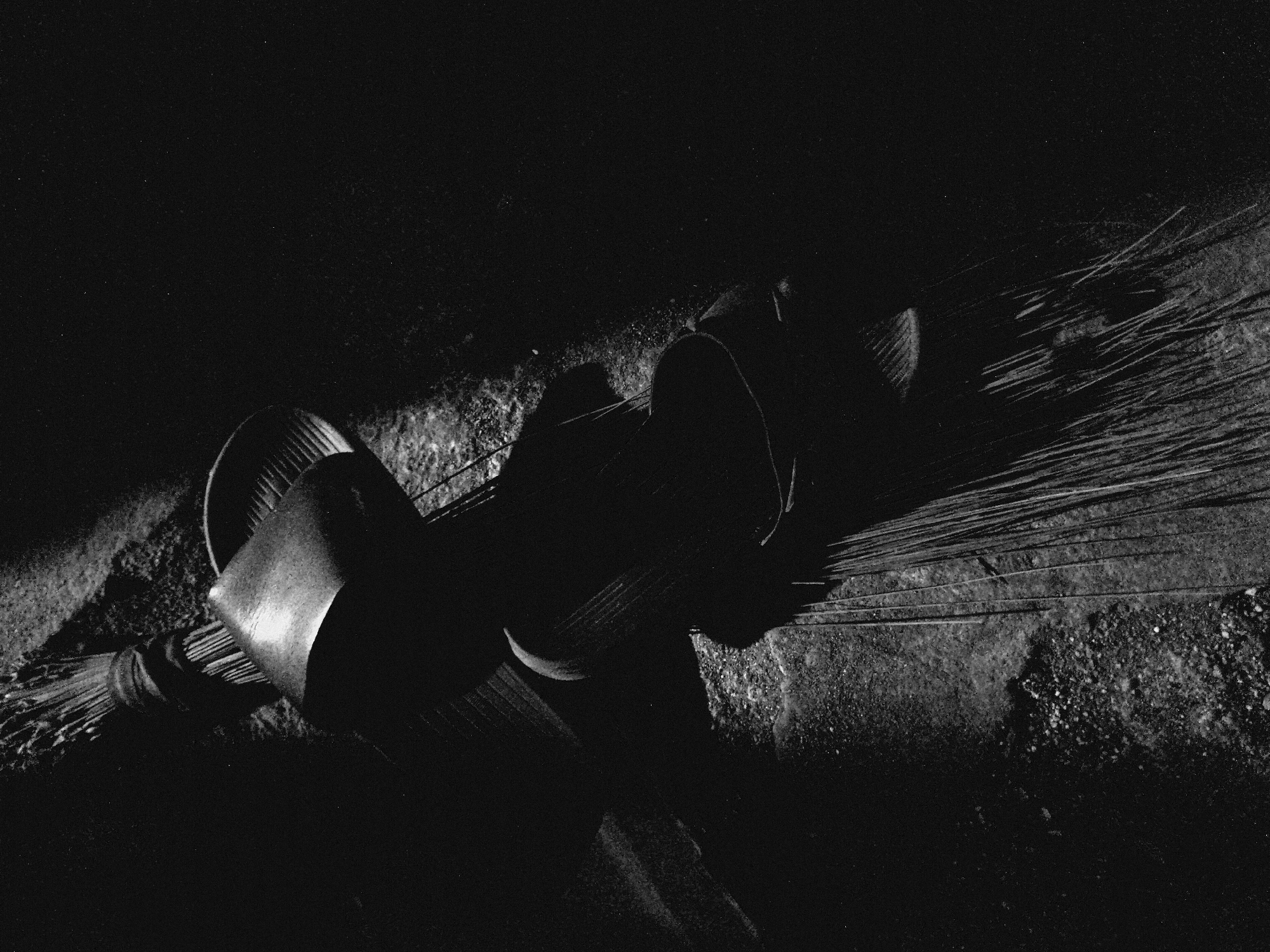 A high-contrast, black and white photograph depicts a broom resting on a textured, rough surface, possibly concrete or stone ground. The composition is diagonal, with the broom's handle and bound head located on the left and center, and its frayed bristles fanning out towards the right side of the frame.
The broom appears to be made of natural fibers, possibly straw or reeds, tightly bound at the handle, which is visible as a wrapped, cylindrical form on the far left. Adjacent to the bound handle, there are broader, curved elements that might be part of the broom's construction or a dustpan positioned alongside it. These elements create strong shadows and highlights due to the dramatic lighting.
The lighting is a prominent feature, characterized by stark chiaroscuro. A focused light source illuminates parts of the broom and the textured ground, casting deep, dark shadows that obscure much of the background and create a sense of depth and mystery. The overall image is dark, with strong whites and grays highlighting textures and forms. The surface beneath the broom shows a gritty, uneven texture.
No specific activity is taking place; the broom appears to be at rest. The time of day is not discernible due to the dramatic and seemingly artificial or localized lighting within a dark environment. There are no people visible, nor any discernible text. The image possesses a visible film grain or digital noise, adding to its stark, raw aesthetic.