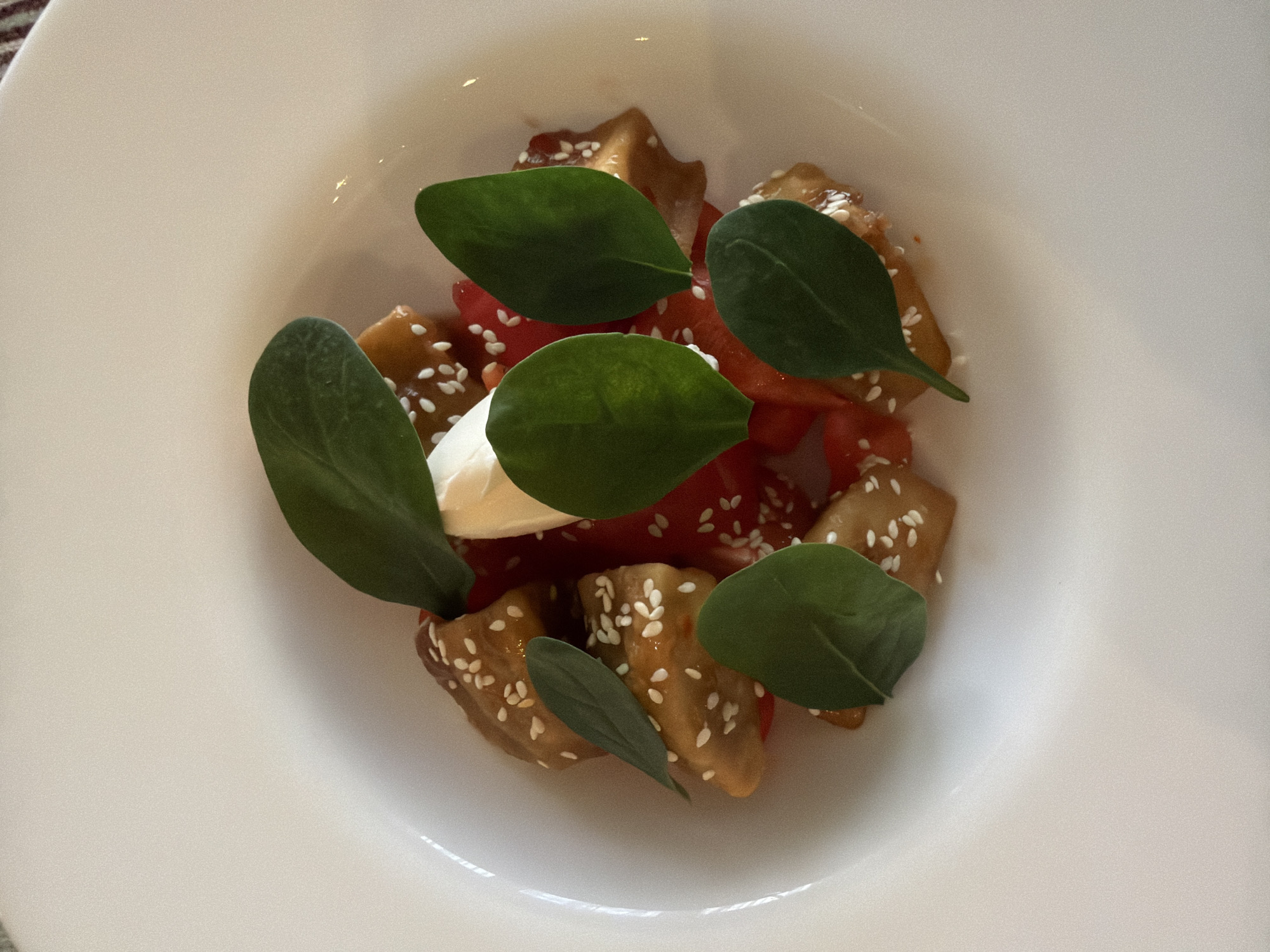 A top-down photographic depiction shows a prepared food dish contained within a white, shallow ceramic bowl. The dish features several irregularly shaped, light brown components, consistent with glazed eggplant pieces, each generously coated with white sesame seeds. These elements are interspersed with diced red material, possibly bell peppers or tomatoes, forming a base or a mixed component. Centrally positioned is a dollop of a white, creamy substance, and the entire composition is garnished with five distinct, dark green leaves, likely spinach or basil, placed decoratively. The bowl rests on an indistinct, light-colored surface. This scene is depicted as being in the city of Chaikovsky, Russia. No people are visible.