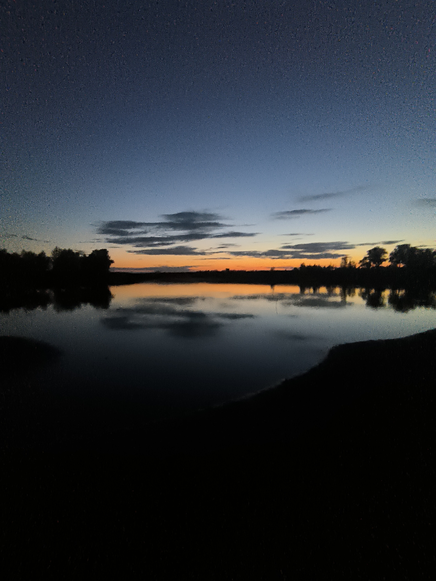 The image depicts a serene landscape at dusk, likely in Zagotzerno, Russia. A calm body of water dominates the foreground, reflecting the fading light of the sky. The sky transitions from a deep twilight blue at the top to warm hues of orange and yellow near the horizon, indicating the setting sun. Scattered clouds are visible, also catching the last rays of sunlight. Silhouetted trees line the far bank of the water, creating a dark, undulating treeline. The overall impression is one of tranquility and the quiet beauty of nature at the end of the day. There are no people or specific objects of note other than the natural elements.