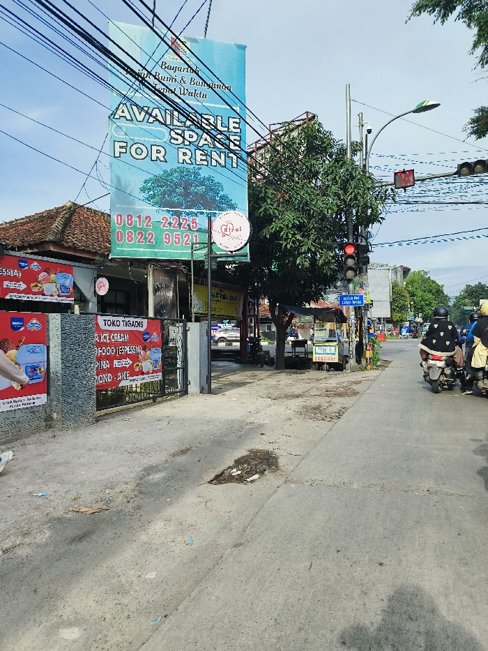 A street scene in Soreang, Indonesia, located at coordinates -7.02 latitude and 107.541 longitude, features a large sign advertising "Available Space For Rent" with phone numbers displayed. The sign is mounted on a pole with a small sign on a wire above it and numerous wires overhead. Two motorcycles are parked on the right side of the street, one with a person wearing a black jacket and helmet. Behind the sign on the left is a building with a sign reading "Toko Tigans Ice Cream, Food, Espresso, Pinda, Rondo, Rice." There are several trees and other businesses along the street. The road surface is gray asphalt with a large pothole in the middle. The sky is a clear blue with a few white clouds.  A traffic light is visible on the right side of the street. There is a street lamp in the background. The scene is taken from a point of view standing on the sidewalk, looking toward the street.  There are several people and cars in the background.  The scene appears to be in daylight.