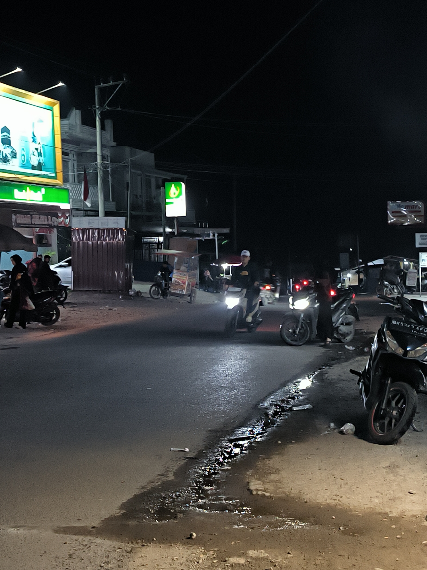 This image depicts a nighttime street scene in Jungke, Indonesia. The foreground shows a paved road with wet patches and debris, indicating recent rain or water accumulation. On the right side of the road, a black motorcycle with a license plate is parked. Further down the road, other motorcycles are visible, some with illuminated headlights, suggesting traffic or people gathered.
In the mid-ground, several individuals are present. Some are standing near motorcycles, while others appear to be walking or operating them. A man is riding a motorcycle in the center of the road. To the left, a group of people are gathered near parked motorcycles and a white car.
The background features buildings, some illuminated by neon signs. One prominent sign on the left displays green and yellow lighting, while another on the right shows a green flame logo. Electrical poles and wires crisscross the dark sky, typical of an urban environment at night. The overall atmosphere is dimly lit, with artificial lights creating shadows and highlighting certain areas of the scene.