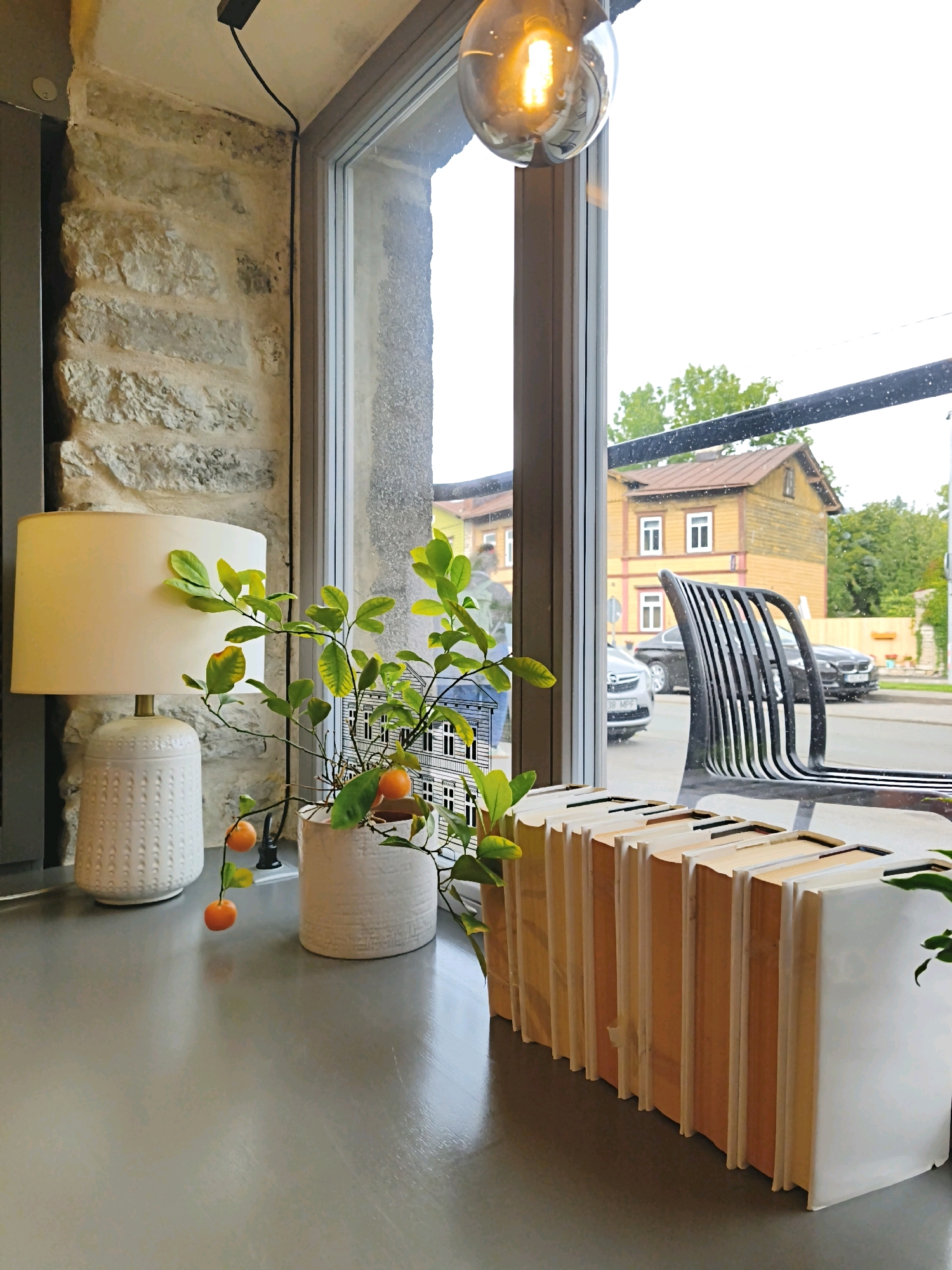 An interior view from a window seat looks out onto a street in Tallinn, Estonia. The foreground features a grey countertop with a white lamp and a potted plant with small orange fruits and green leaves. To the right, a row of white and wood-colored objects resembling books is arranged on the countertop. Beyond the window, a black metal chair is visible. The street outside shows parked cars and two colorful wooden houses. The sky is overcast.