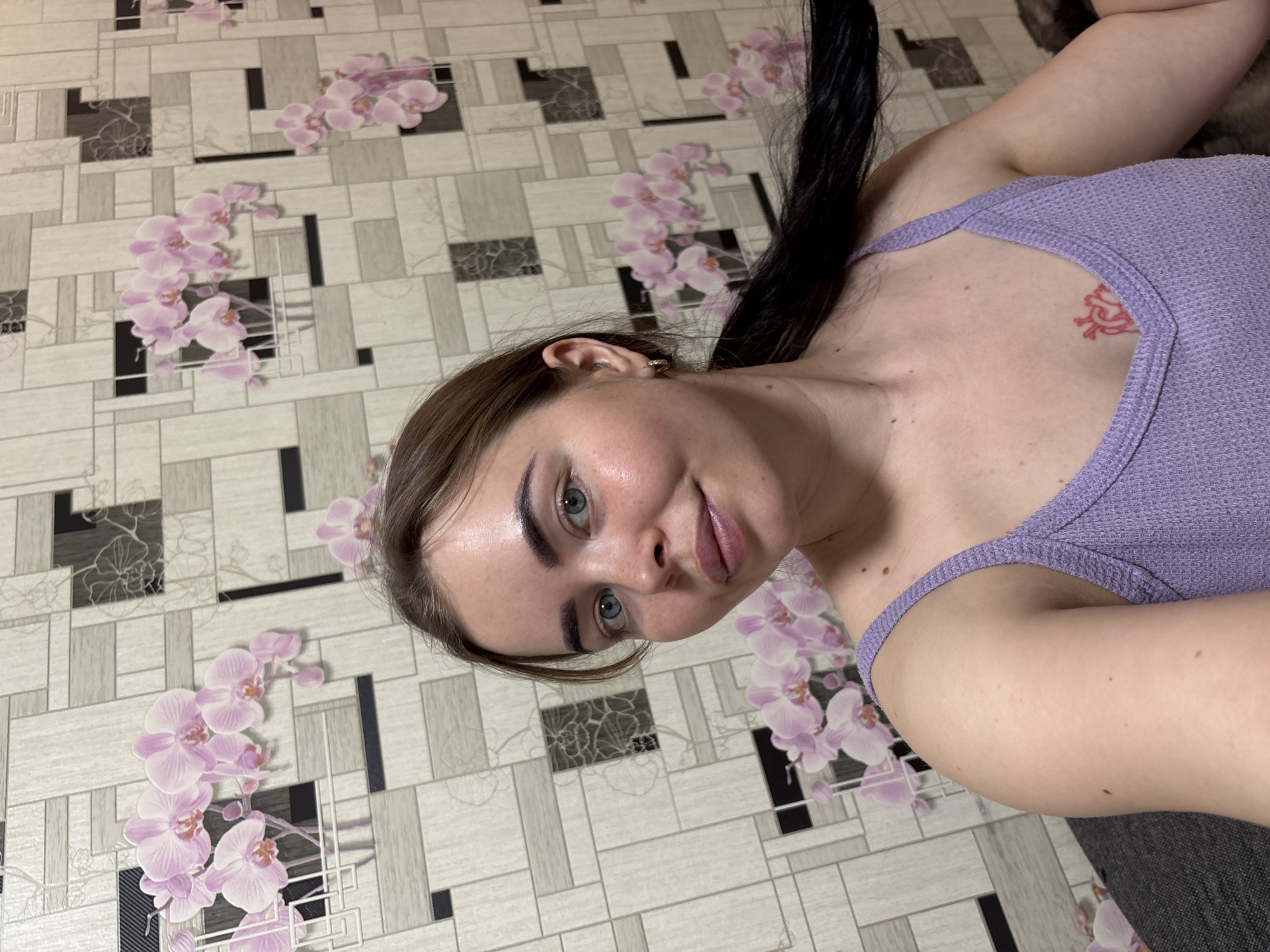 A woman with fair skin and blue eyes is seen in a selfie-style photograph. She is wearing a lilac-colored top and has dark brown hair with some black strands. There's a reddish tattoo on her upper chest. The background appears to be wallpaper with a geometric pattern and floral designs featuring pink orchids. The setting is indoors. The image's orientation is rotated, giving the impression that the subject is lying down.
While it's impossible to confirm the location in the city of Izhevsk, Russia, just from the image alone, the provided information suggests that the location may be pertinent.